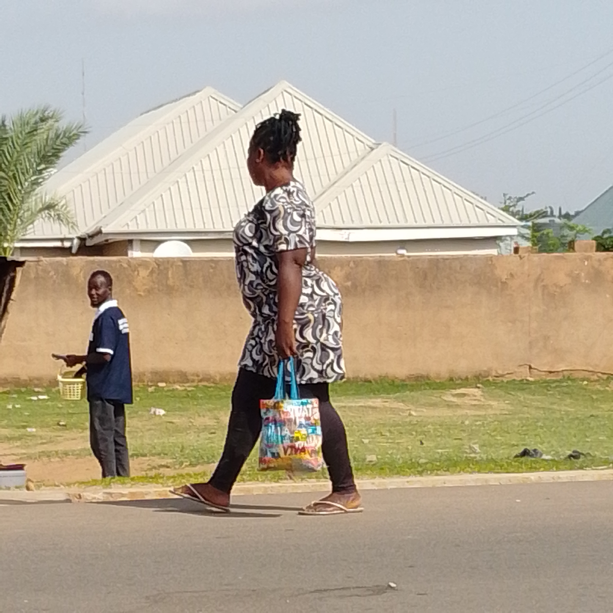 In Tempure, Nigeria, a woman with dark hair pulled back in a bun walks from right to left across a paved road. She is wearing a short-sleeved, patterned dress in shades of black, white, and gray, and black leggings. In her right hand, she carries a multi-colored tote bag with the words "VIVAT" and "VIVA" visible on it. On her feet, she wears brown flip-flops. To her left, a man in a dark blue shirt with white stripes on the sleeves and collar, and dark pants, stands facing the camera. He is holding a small object in his right hand and a woven basket in his left. Behind the people, a low, textured beige wall runs horizontally, with a white-gabled building with visible metal roofing behind it. A palm tree is visible on the far left of the image. The ground consists of patchy green grass and the paved road. The sky is light blue and overcast.