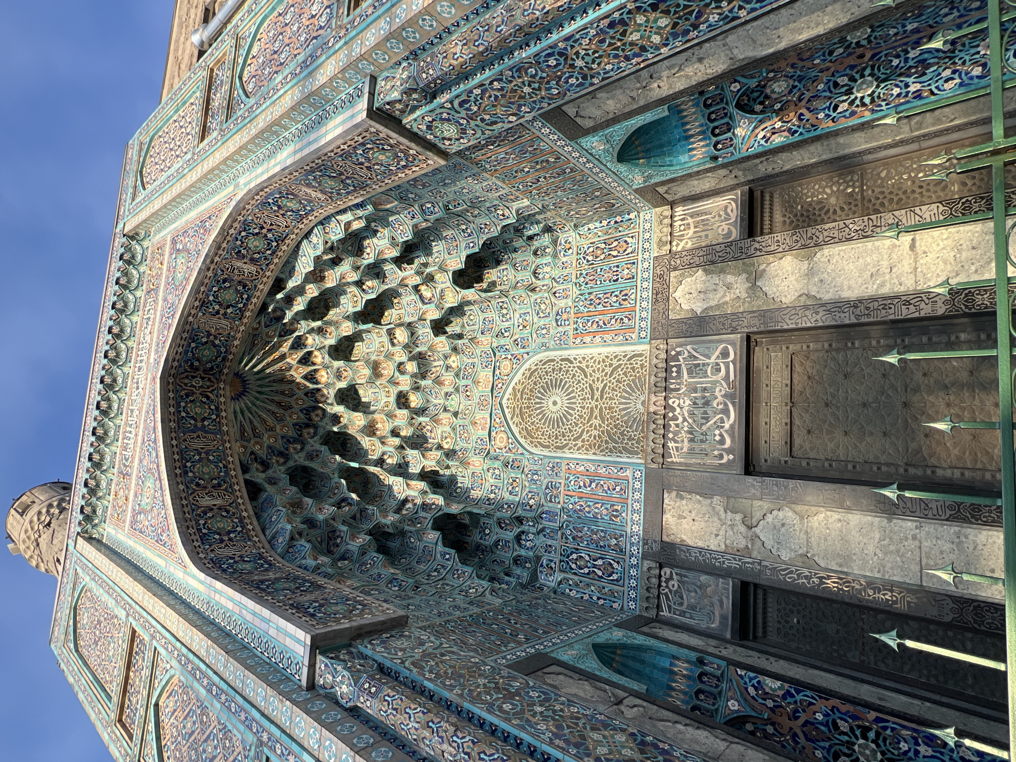 This is a low-angle, eye-level photograph of the Saint Petersburg Mosque in Russia. The architecture features detailed tilework in shades of blue, green, and brown. The entrance is arched, and there is Arabic calligraphy inscribed on the walls. The photo also captures a minaret against a clear blue sky.