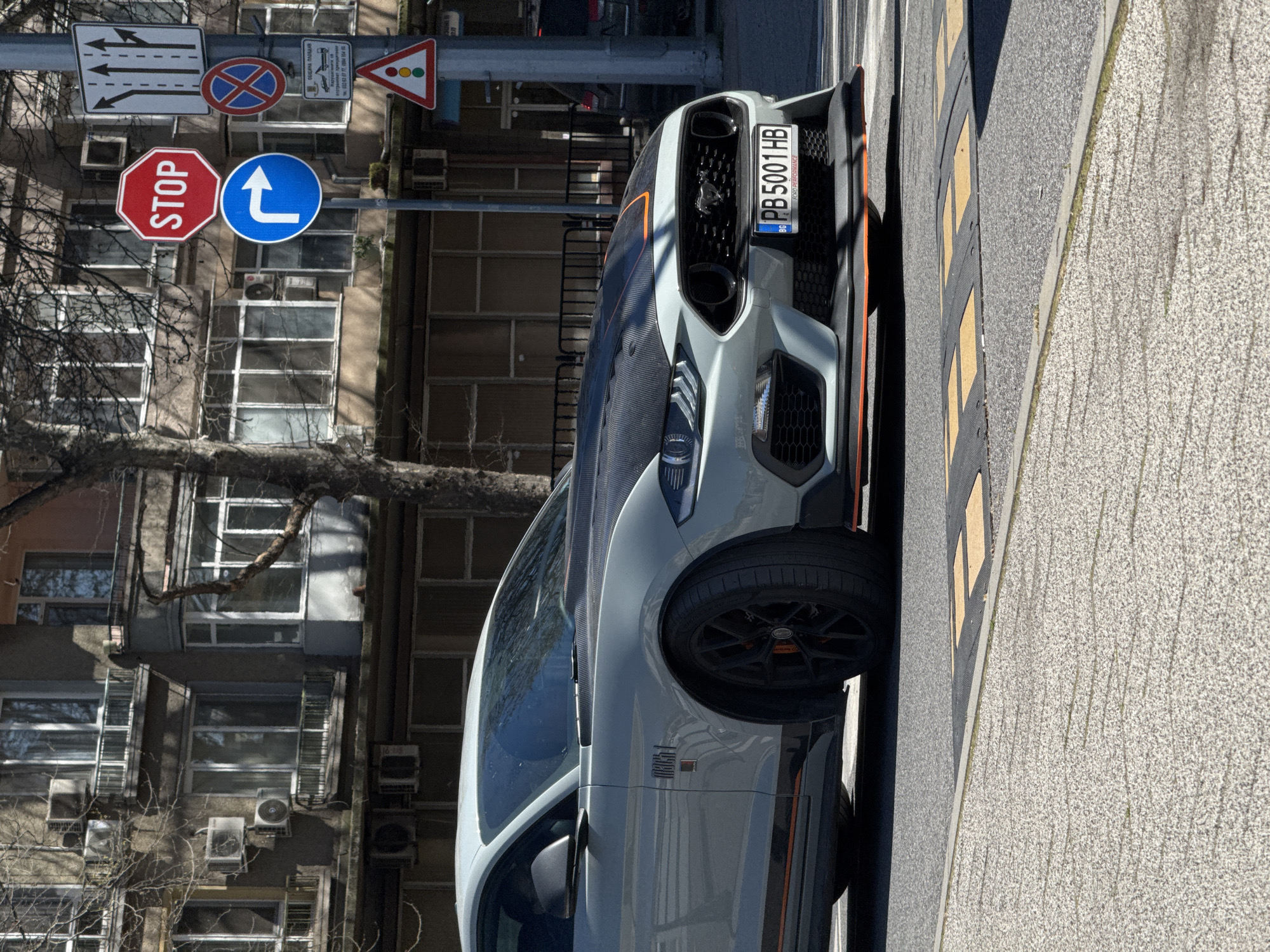 The image shows a gray Ford Mustang Mach 1 parked on a street in Plovdiv, Bulgaria. The car is positioned close to the curb, partially on a section with yellow and black speed bumps. The car's front is facing the top right of the frame. The license plate reads "PB 5001 HB". In the background, there are apartment buildings with multiple windows, and some trees with bare branches. There are various road signs visible in the upper left corner of the image, including a stop sign, a blue sign with a white arrow indicating a left turn, and others.