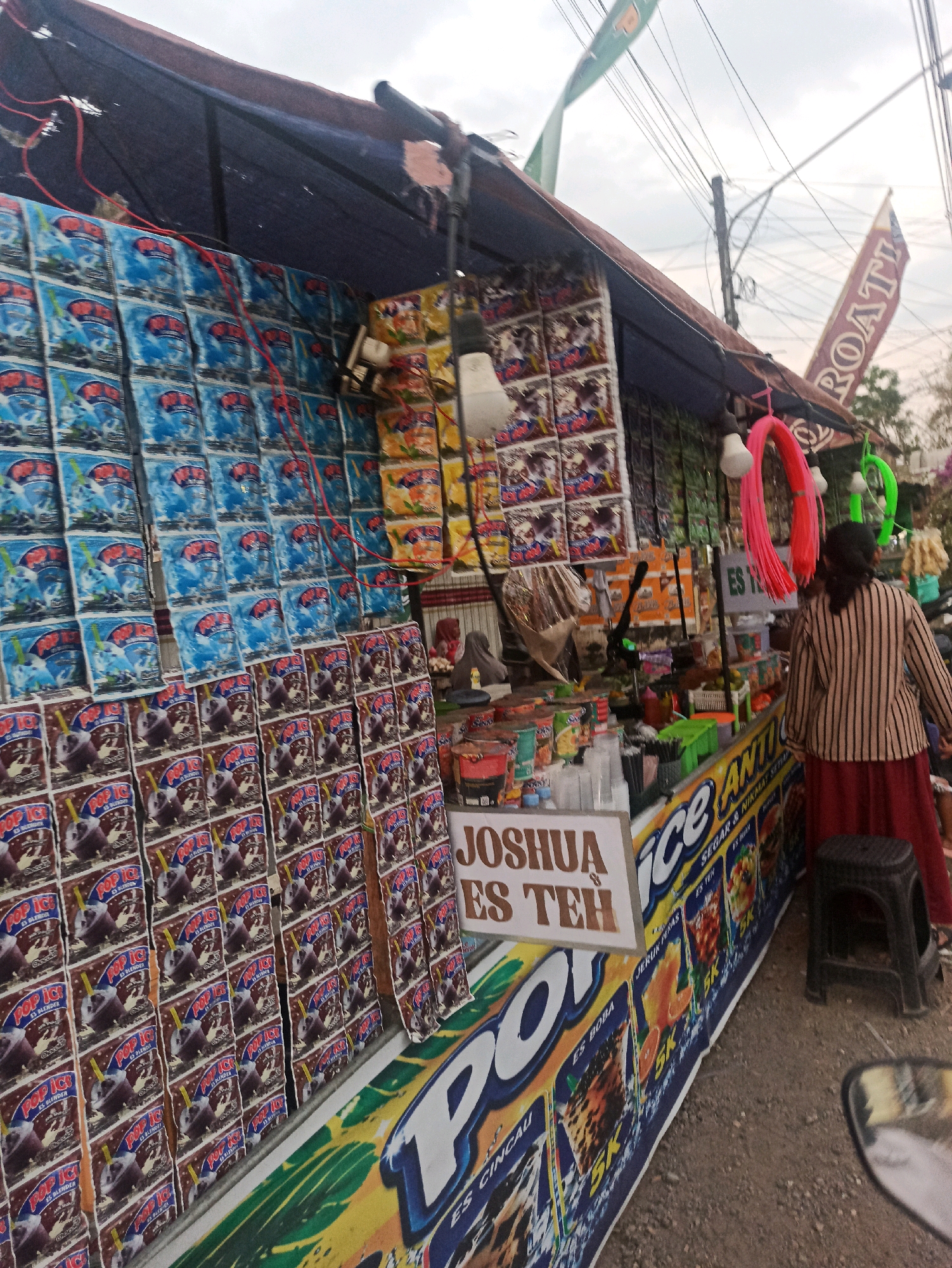The image depicts a street-side stall in Tlogosari, Indonesia, offering a variety of packaged drinks and snacks. The stall is covered by a blue and maroon awning, with red electrical wires strung across the top. The front of the stall is adorned with rows of brightly colored product packaging, predominantly featuring "Pop Ice" branding with images of frozen drinks. A prominent sign reads "JOSHUA ES TEH," indicating the stall's name and specialty. To the right, a woman wearing a striped shirt and a red skirt stands near the stall, facing away from the camera. Several colorful hanging lights and decorative ropes, pink and green, are visible above the counter. The ground is a dirt and gravel surface. The background shows a cloudy sky and utility wires.
