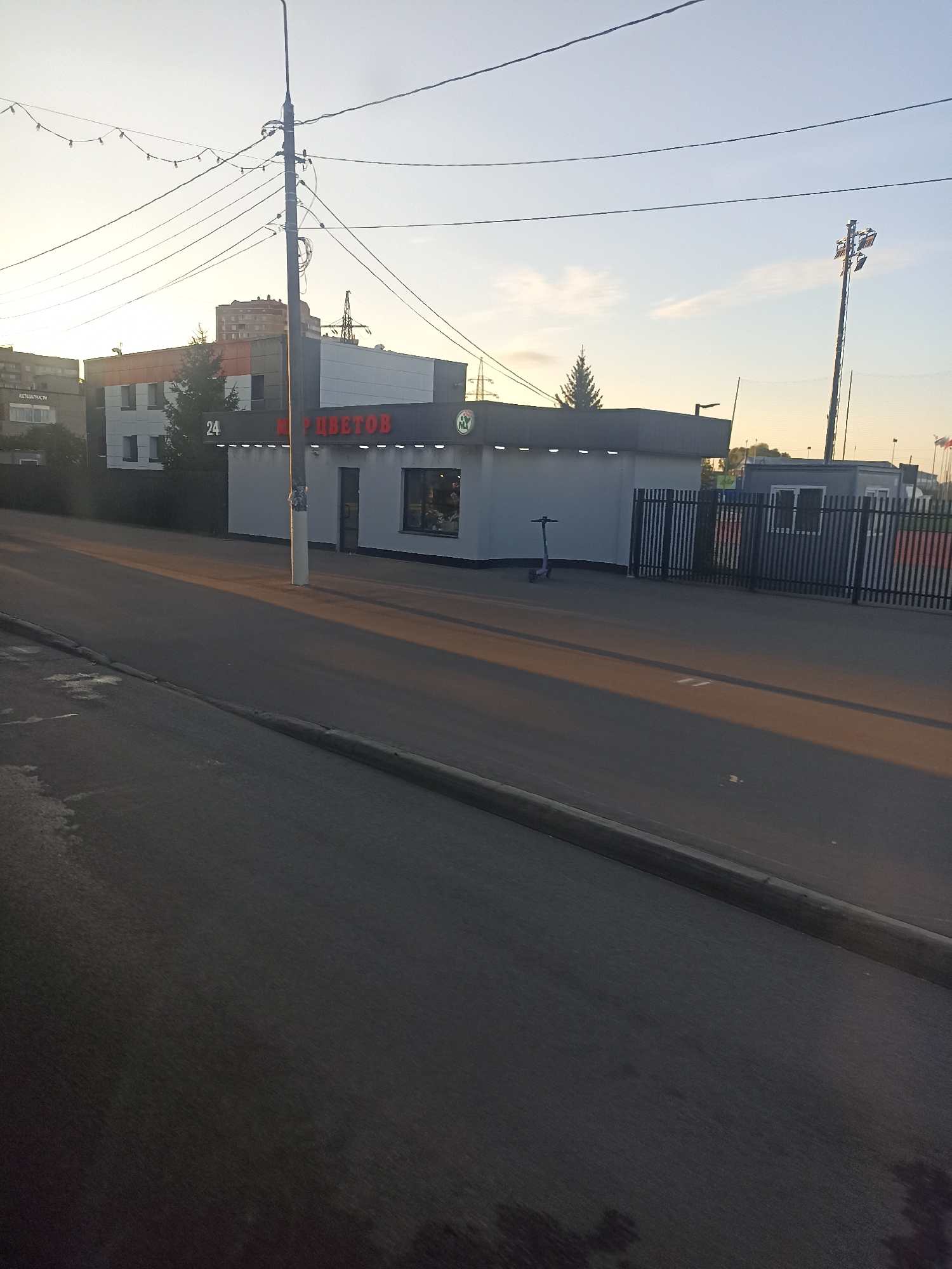 The image shows a street scene in Chekhov, Russia, during daylight. In the foreground, a road with a curb is visible, bathed in the warm light of the sun. To the right of the road, there is a sidewalk and a small building that appears to be a shop. This building has a sign with red Cyrillic lettering that reads "МИР ЦВЕТОВ" (World of Flowers) and a "24" indicating it is open 24 hours. There is also a green logo with a star on the building. Next to the shop is a black metal fence. A purple electric scooter is parked on the sidewalk in front of the shop. Further in the background, other buildings, including a multi-story residential building, are visible, along with power lines and streetlights. The sky is clear with a few wispy clouds. The overall impression is of a typical urban streetscape.