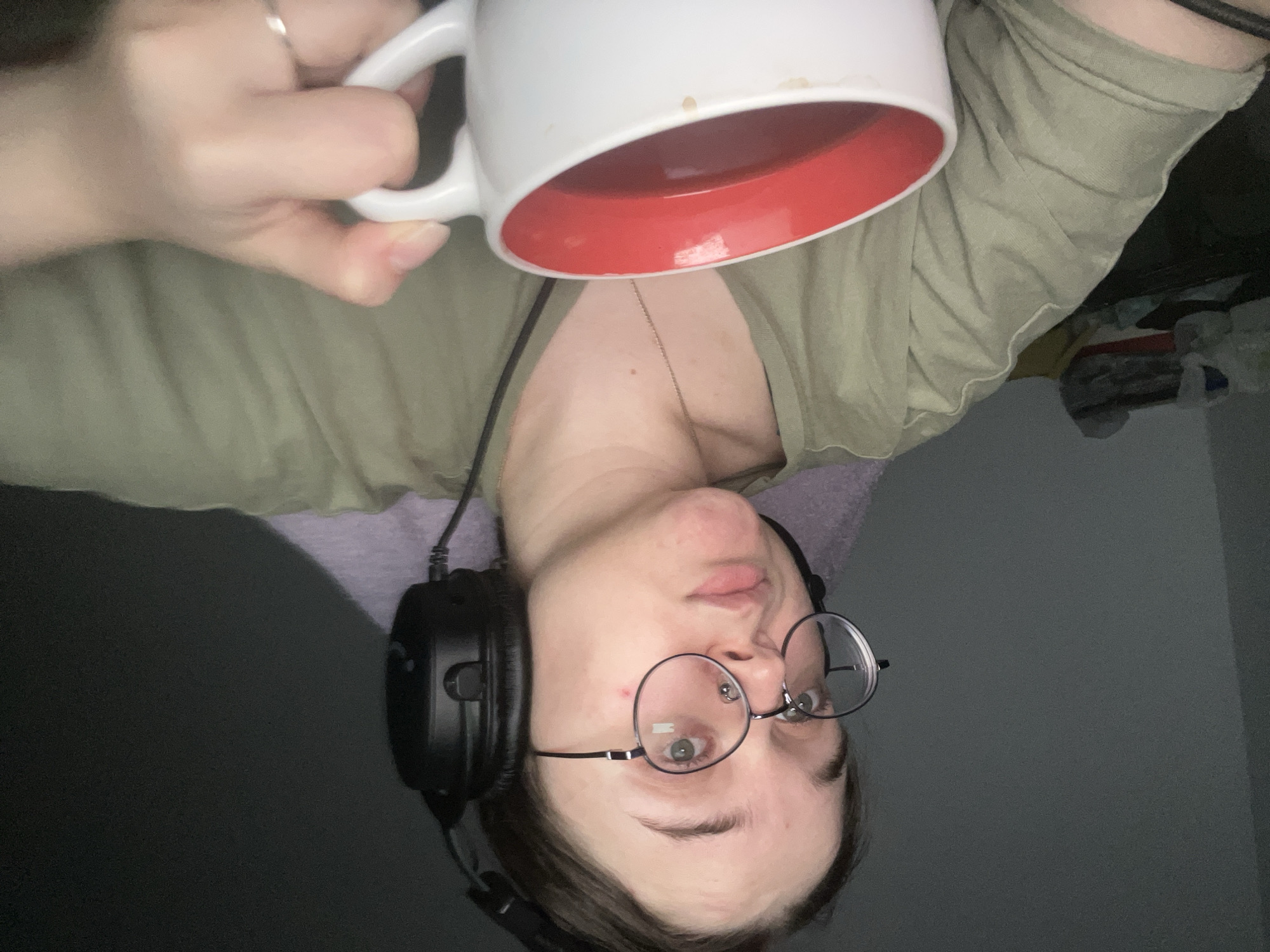 An inverted, close-up shot features a person with short dark hair, wearing round-framed glasses and a nose piercing, appearing to look upwards towards the camera. The individual wears a light olive-green top and has black over-ear headphones draped around their neck. A thin necklace is visible on their chest. They are holding a white mug with a vibrant red interior, filled with a dark liquid, likely coffee or tea. Some dried residue is present on the outside rim of the mug. A silver ring adorns their ring finger.
The person's skin is fair, and a small red mark is visible on their cheek. Their mouth is slightly open, and their gaze is directed towards the lens. The background is dark gray/black on one side and a lighter, possibly fabric texture on the other, suggesting an indoor setting with low or artificial lighting. Indistinct items are faintly visible in the far background on the right. The overall impression is a casual, relaxed moment. The image is captured in Pushchino, Russia. No text is visible in the frame.