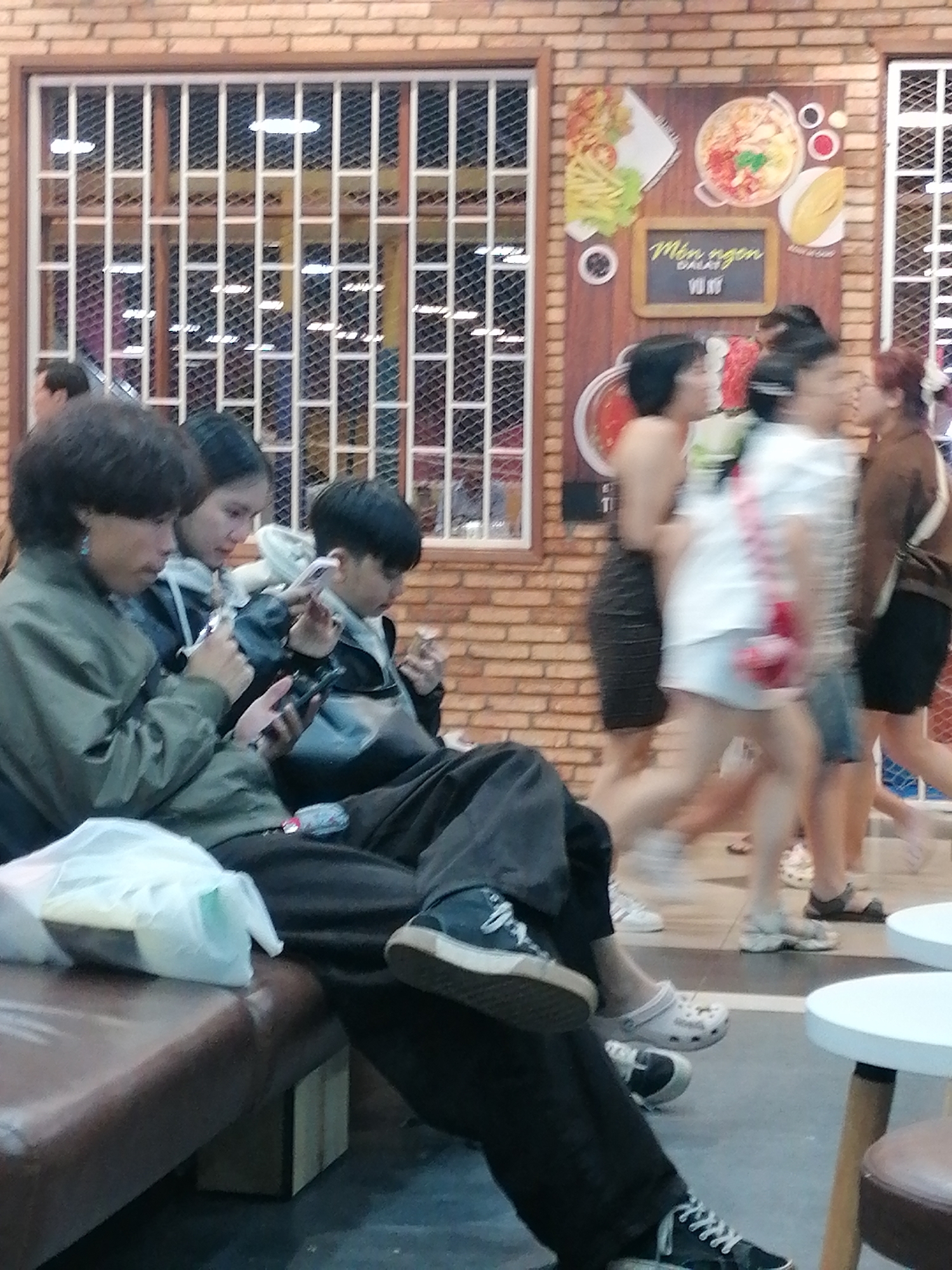 The image depicts a street scene in Da Lat, Vietnam, featuring several individuals and a brick wall with a decorative mural and a caged window. In the foreground, three young people are seated, each engrossed in their mobile phones. The person on the left, wearing a green jacket, has dark, medium-length hair and is looking down at their phone. To their right, a young woman with dark hair in a hoodie also focuses on her phone. Next to her, a young person with short dark hair is using their phone, and their left hand appears to be holding a small object.
In the background, several other people are visible, some walking and others standing. One woman in a black dress is seen from the side, and another in a white top and denim shorts is walking, appearing somewhat blurred. Further to the right, a person in a brown top is also walking.
The environment includes a brick wall with a large mural depicting food items, including a bowl of noodles and other dishes, with Vietnamese text that reads "Món ngon DALAT" (Delicious dishes of Da Lat) and "Vui Vẻ" (Joyful). A white-framed, barred window is set into the brick wall above the mural. A brown leather bench is visible in the lower left, with a white plastic bag on it. Two white circular tables with wooden legs are present in the lower right of the frame. The flooring appears to be a light-colored tiled surface. The overall impression is of a casual, public space, possibly a cafe or food stall, in Da Lat.