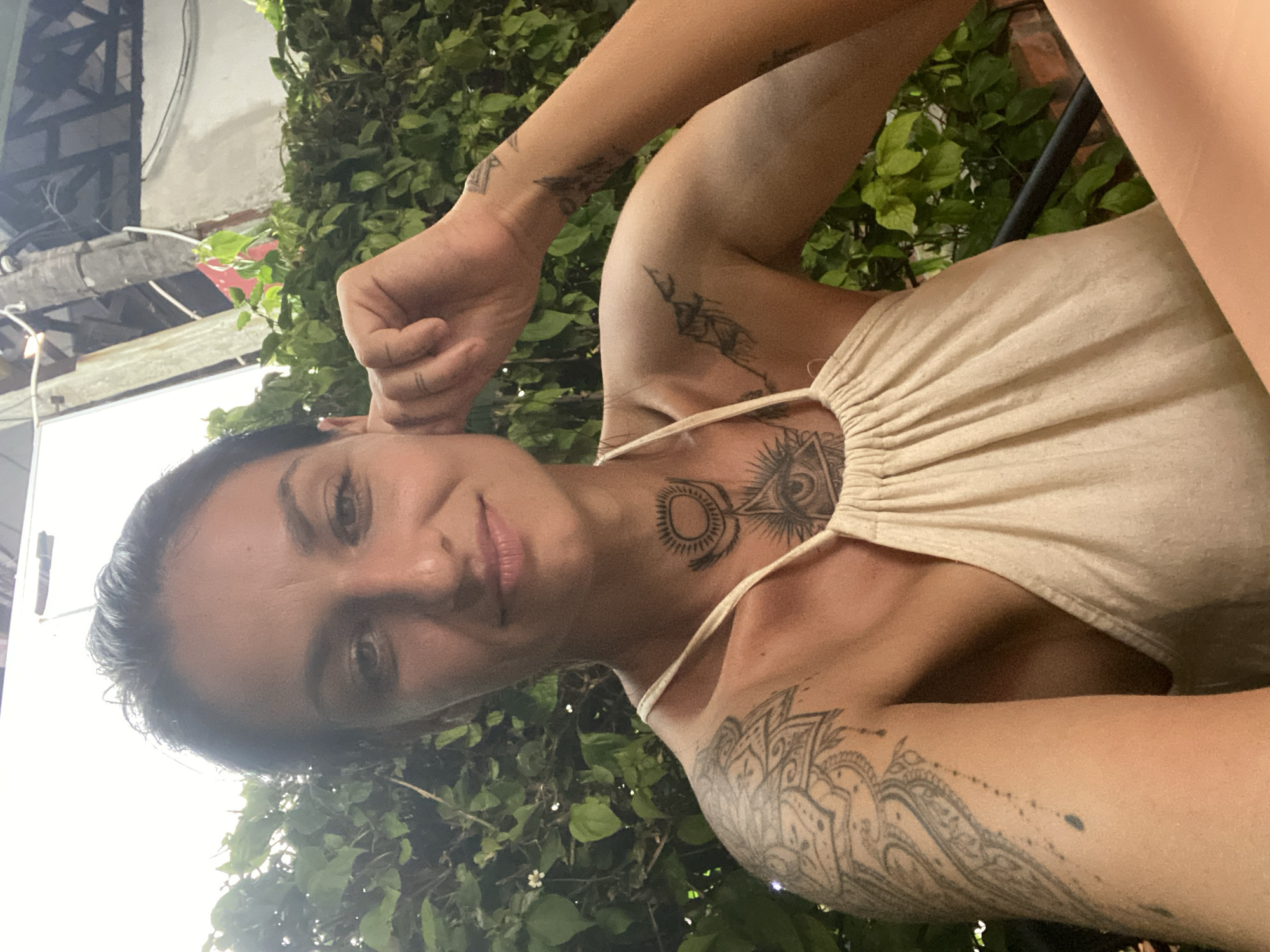 A woman with dark hair and tattoos on her arms and chest is sitting at a table outdoors, with a lush green hedge behind her. She is wearing a light-colored, halter-neck top. Her right hand is resting on her cheek as she smiles at the camera. The scene appears to be in a casual outdoor dining area, possibly a restaurant or cafe in Phu Thuy Ward, Vietnam, as indicated by the presence of dense foliage and outdoor seating. The lighting suggests it might be late afternoon or early evening.