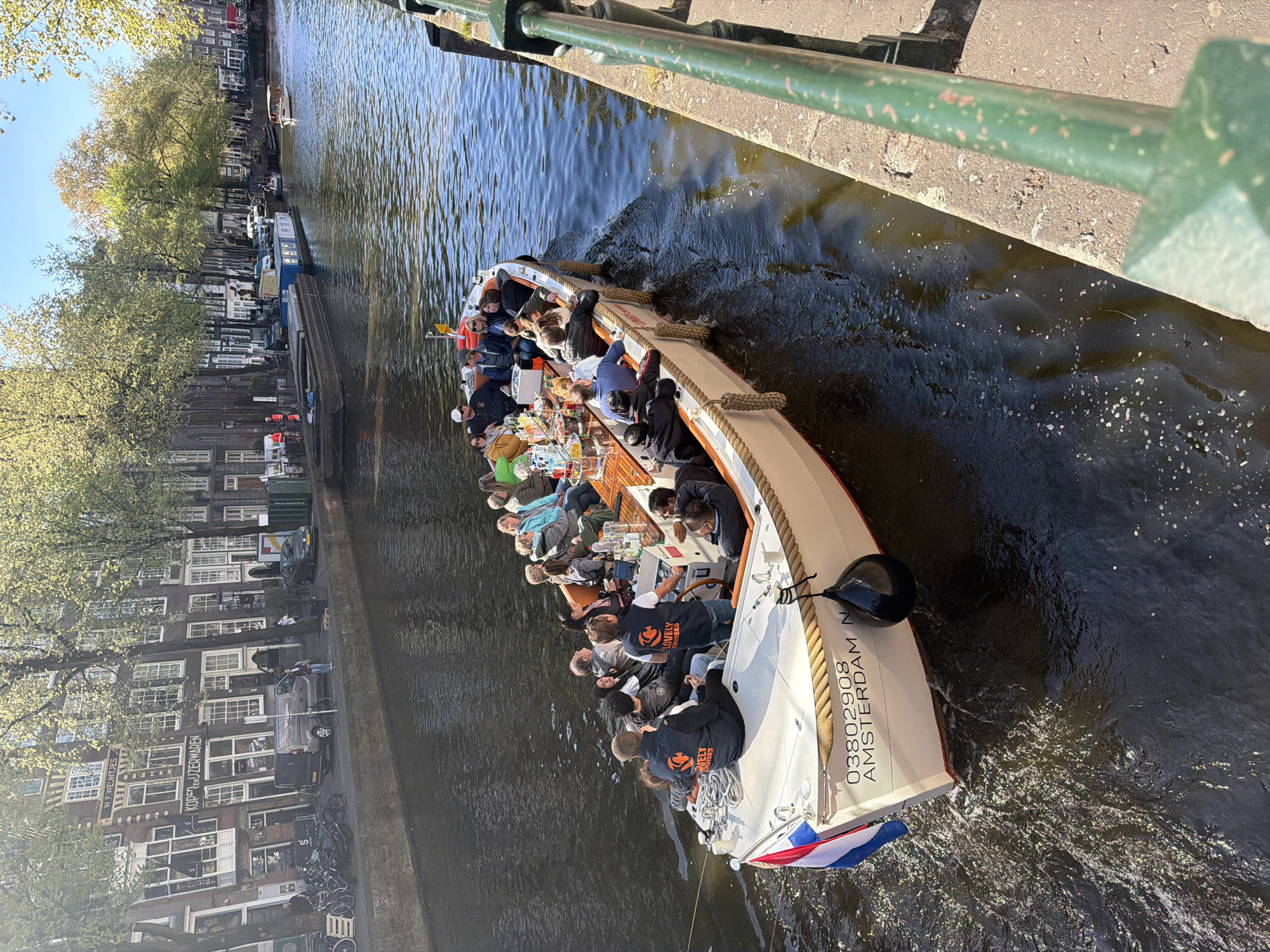 A canal boat filled with people is cruising along a waterway in Amsterdam, Netherlands, on a bright, sunny day. The image is taken from an elevated perspective, looking down from a bridge, with a green metal railing visible in the foreground.
The white and brown canal boat is packed with passengers, many of whom are seated at tables enjoying drinks and food. Some individuals on the boat are wearing dark jackets with an orange logo and the text "LOVELY CRUISES." On the stern of the boat, the Dutch flag is visible, along with the registration number "03802908 AMSTERDAM N." The water around the boat shows ripples and sunlight reflections, indicating movement.
Along the left bank of the canal, a row of typical Amsterdam canal houses lines the street. These historic buildings have multiple windows and distinctive architecture. Lush green trees with light spring foliage stand in front of the buildings. On the canal bank, bicycles are parked, and cars are visible on the street. Some visible text on the buildings includes "W.F. WEIJLATES" and "KOPERWERVEN" (partially obscured, likely "Koperwerken"). Another, smaller boat is visible further down the canal.
The scene depicts a leisurely canal tour under clear skies, with the people on board appearing relaxed and engaged in the experience.