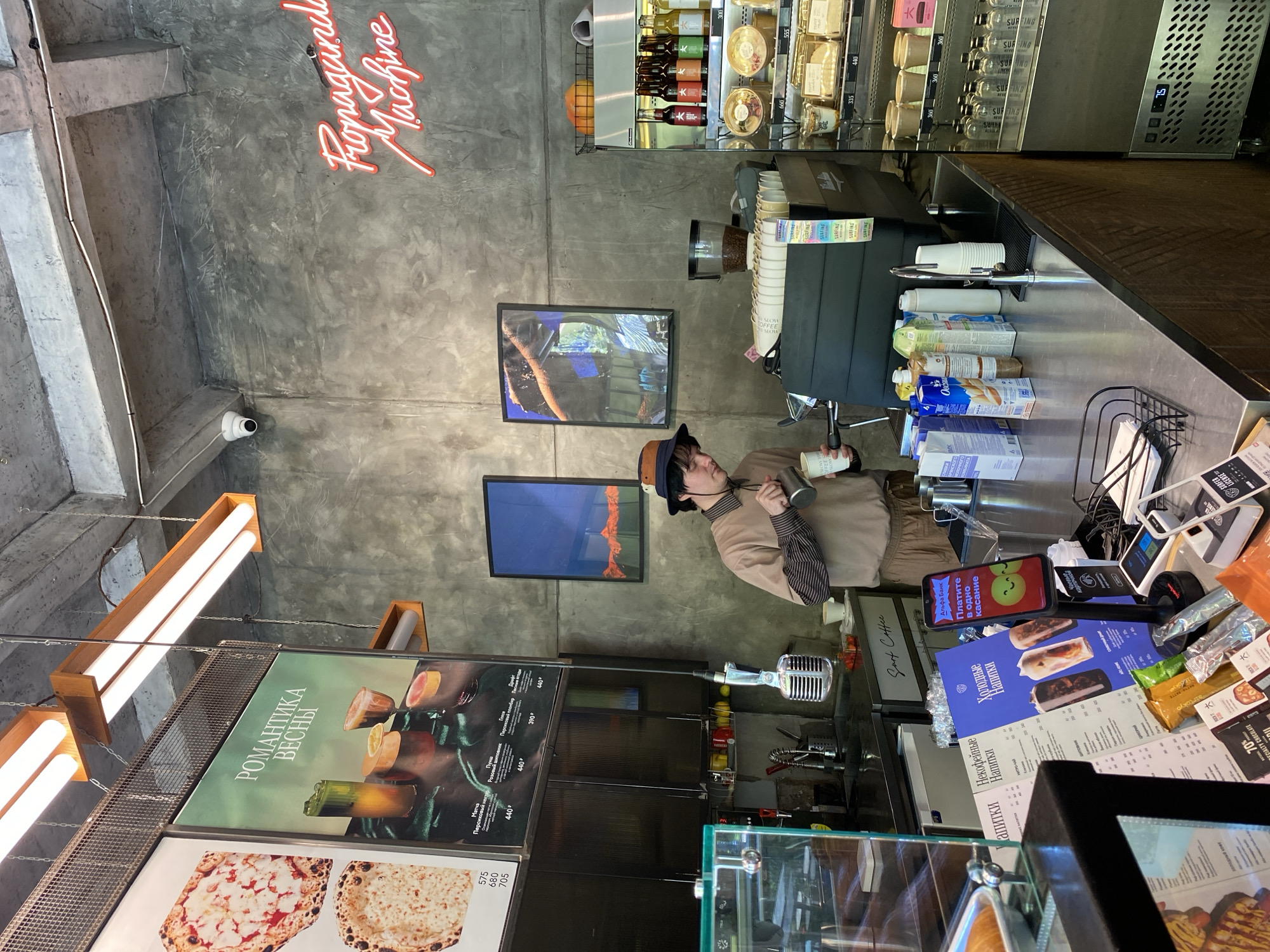 This image captures a scene inside Surf Coffee in Sochi, Russia. The walls have a raw concrete finish, and a red neon sign reading "Propaganda Machine" is visible on one wall. There are two framed pictures hanging on the wall. A man is standing behind a counter, wearing a hat and holding a coffee cup. The counter is made of stainless steel and contains various items, including coffee supplies, milk cartons, and a point-of-sale system with a phone displaying "Альфа Банк" (Alfa Bank) and "Платите в одно касание" (Pay in One Touch) in Russian. A menu board and display case with food items are visible in the lower part of the image. Above the menu board, a rectangular light fixture illuminates the scene.