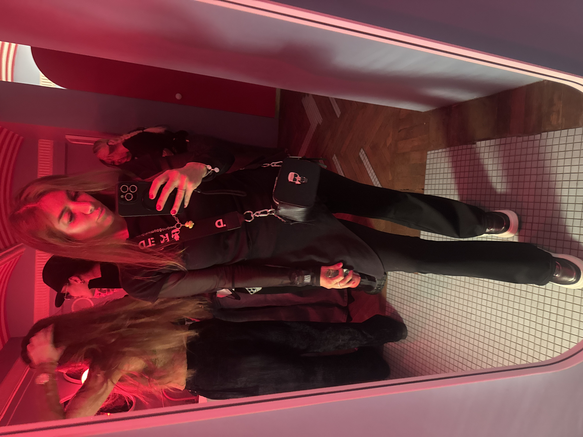 A photograph shows two people reflected in a mirror inside a room with a red light. One person has long, reddish-blonde hair and is wearing a black outfit, holding a black smartphone as if taking a selfie or recording a video. The phone appears to have three camera lenses. The other person wears a black baseball cap and stands behind. A small black purse with a silver skull detail hangs from the first person's waist. The floor visible in the reflection is tiled in a white grid pattern, while adjacent areas feature herringbone-patterned wooden floors. The surrounding structure is bordered with a light blue frame. It is likely a photograph taken inside a restaurant or other public space in Moscow, Russia.