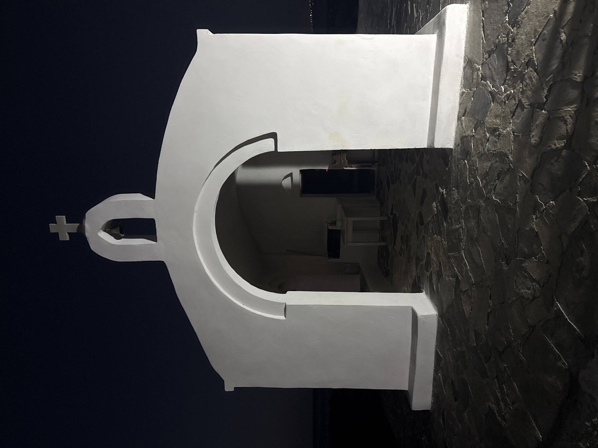 The image shows a small, white, chapel-like structure with a cross on top, illuminated against a dark blue sky. The chapel is built into a rough, stone wall. The structure has a curved archway leading into a dark interior, where a few small white objects are visible. The lighting suggests it is nighttime, and the overall mood is serene and perhaps spiritual. The location cues, combined with the architectural style, point to a Mediterranean setting, consistent with Georgioupoli, Greece. No text is visible in the image.
