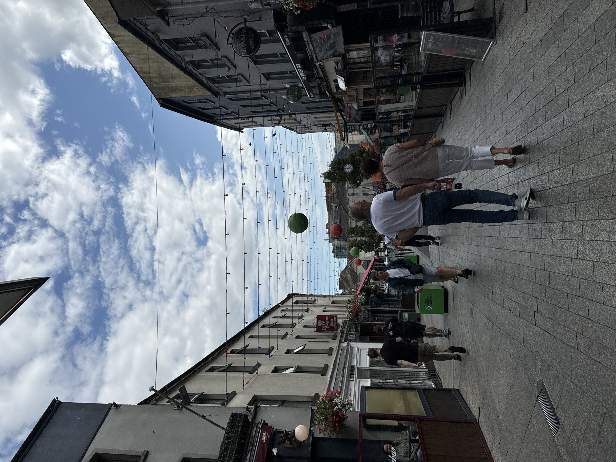 The image shows a pedestrianized street in Waterford, Ireland. The street is lined with buildings on both sides, many of which appear to be shops or pubs. Strings of colorful lights and large green and red spheres are suspended overhead, suggesting a festive atmosphere. Several people are walking along the street. In the foreground, a couple walks away from the camera, with the man wearing a white polo shirt and blue jeans, and the woman in a light-colored top and white trousers. Further up the street, other individuals can be seen walking in various directions. The paving stones are laid in a pattern, and there are green planters on the sidewalks. The sky is partly cloudy, with blue patches visible. A sign for "The Market Bar" is visible on the right side of the street. The overall impression is that of a lively, urban commercial area.