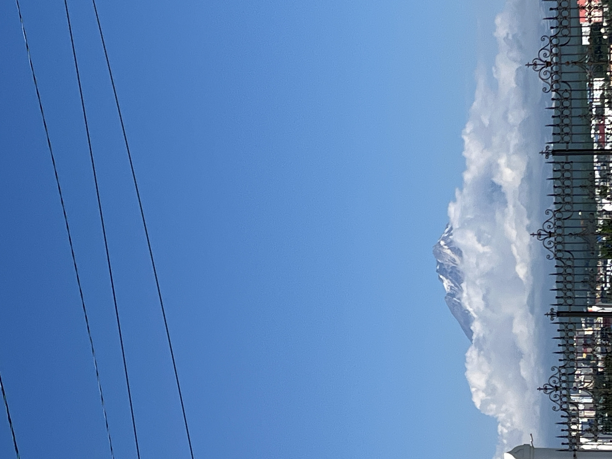 In Petropavlovsk-Kamchatsky, Russia, a mountain peak with snow-capped slopes pierces through a layer of white clouds under a clear blue sky. Three parallel power lines are strung across the top of the frame. In the foreground, a decorative black metal fence runs horizontally, with a white pillar visible on the left. Behind the fence, a cityscape with buildings and trees can be seen.