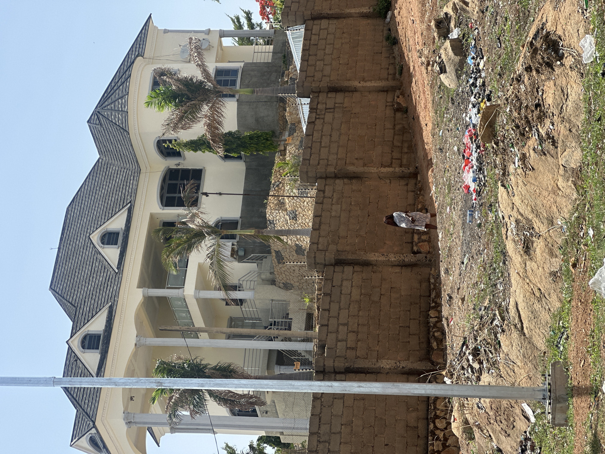 The image captures a three-story building in Jos, Nigeria, characterized by a beige exterior, a dark gray tiled roof, and white columns. Palm trees are visible in front of the building. Adjacent to a brick wall, a slope shows signs of garbage and waste scattered across the terrain. A person is standing near the wall. A metal pole runs vertically across the bottom left of the frame. The sky is clear and blue.
