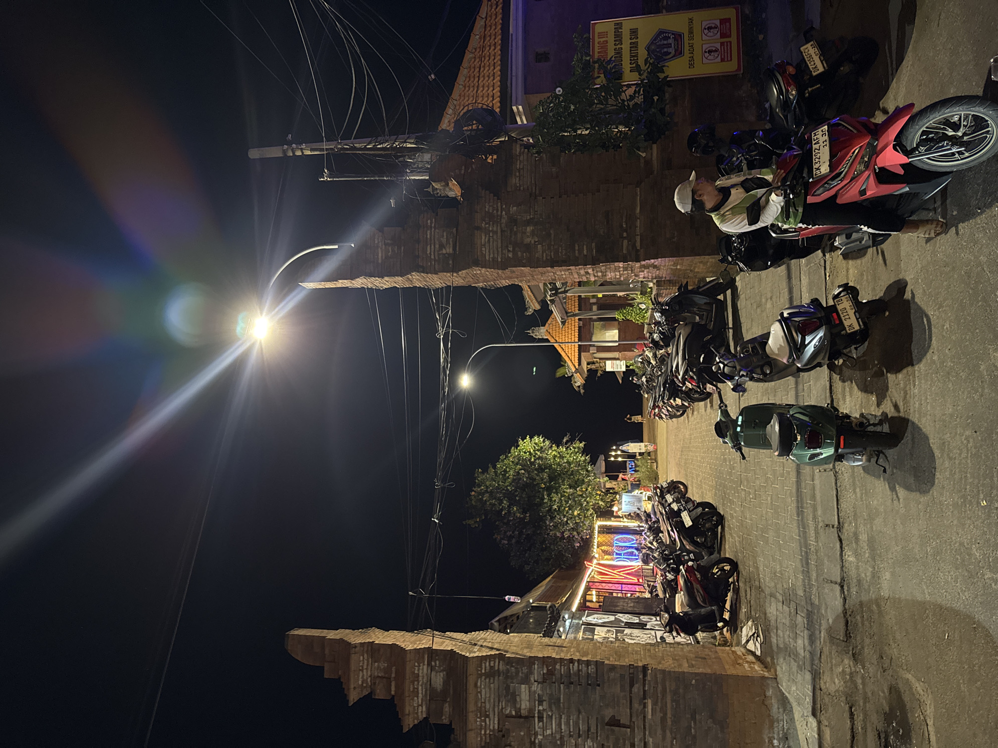 This is a night shot of a street in Kuta, Indonesia, with traditional Balinese brick archways framing the view. A street lamp with lens flare illuminates the scene. Numerous scooters and motorcycles are parked along the paved street, which is lined with buildings featuring thatched roofs. One building has neon signs that read "RESO" and "V." A man wearing a white cap, green and white shirt, and black pants is seated on a red scooter. Another person is visible walking in the distance. A yellow sign with Indonesian text is mounted on a pole. The environment appears to be a busy commercial area at night, likely a tourist destination.