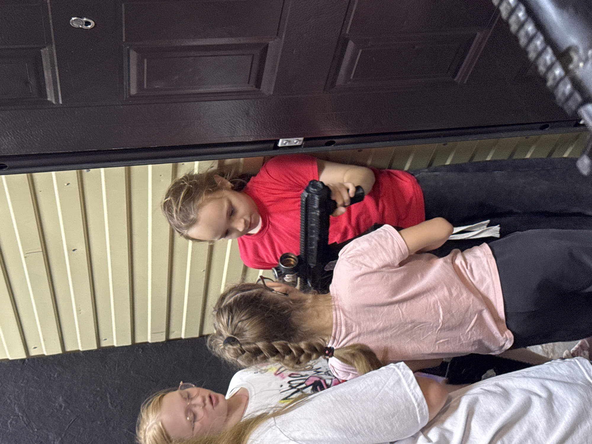 In Perm, Russia, two young girls are positioned near a dark brown door with a peephole and a white horizontal siding wall. The girl on the right, wearing a red t-shirt and dark pants, is holding a black toy gun and appears to be looking at something intently. The girl on the left, wearing a pink t-shirt and dark pants, has her back to the camera and is also looking in the same direction as the other girl. Her hair is styled in a thick braid. Another girl, partially visible on the far left and wearing a white t-shirt with colorful graphics, is looking towards the girl with the braid. The scene suggests a playful interaction, possibly a game, taking place outdoors or in a transitional area like a doorway.