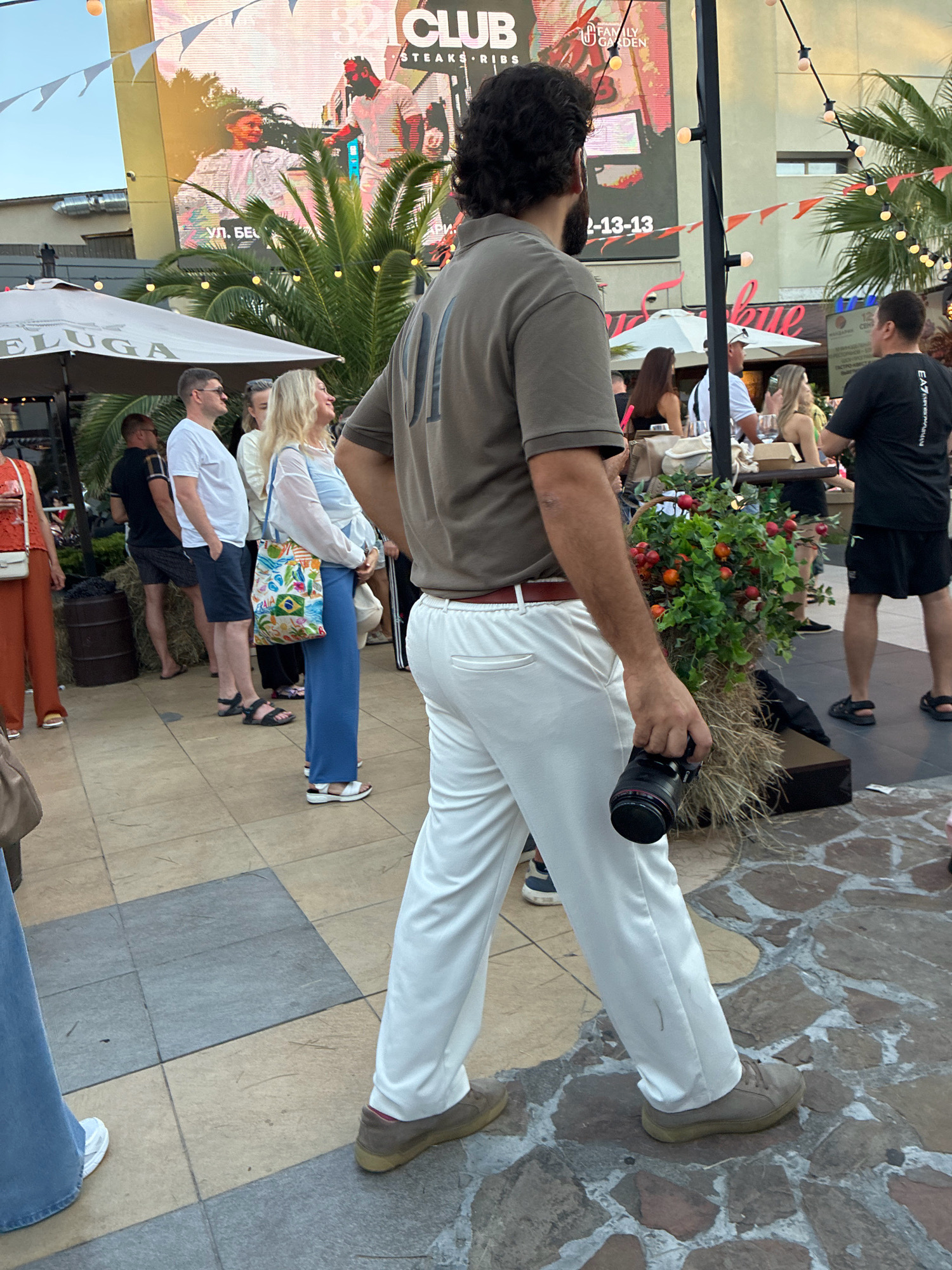 The image depicts an outdoor scene, likely in Sochi, Russia, given the presence of palm trees and a bright, sunny atmosphere. A man with dark, curly hair and a beard, wearing a khaki polo shirt and white trousers, is walking to the right, holding a black camera in his right hand. He is the primary subject in the foreground.
Behind him, a group of people are milling about. To the left, a woman in a white top and blue pants is looking towards the camera, carrying a colorful tote bag. To her left, a man in a white t-shirt and dark shorts stands with his hands clasped. Further back, other individuals are visible in casual attire, some standing, others seated at tables.
The environment suggests a public area, possibly a promenade or an outdoor dining/shopping district. Large umbrellas are visible, one with "BELUGA" printed on it. A large LED screen displays an advertisement featuring people and text. Decorative elements like string lights and potted plants with red berries add to the festive ambiance. The ground is paved with square tiles and irregular stone patterns. The overall impression is a lively gathering on a pleasant day.
