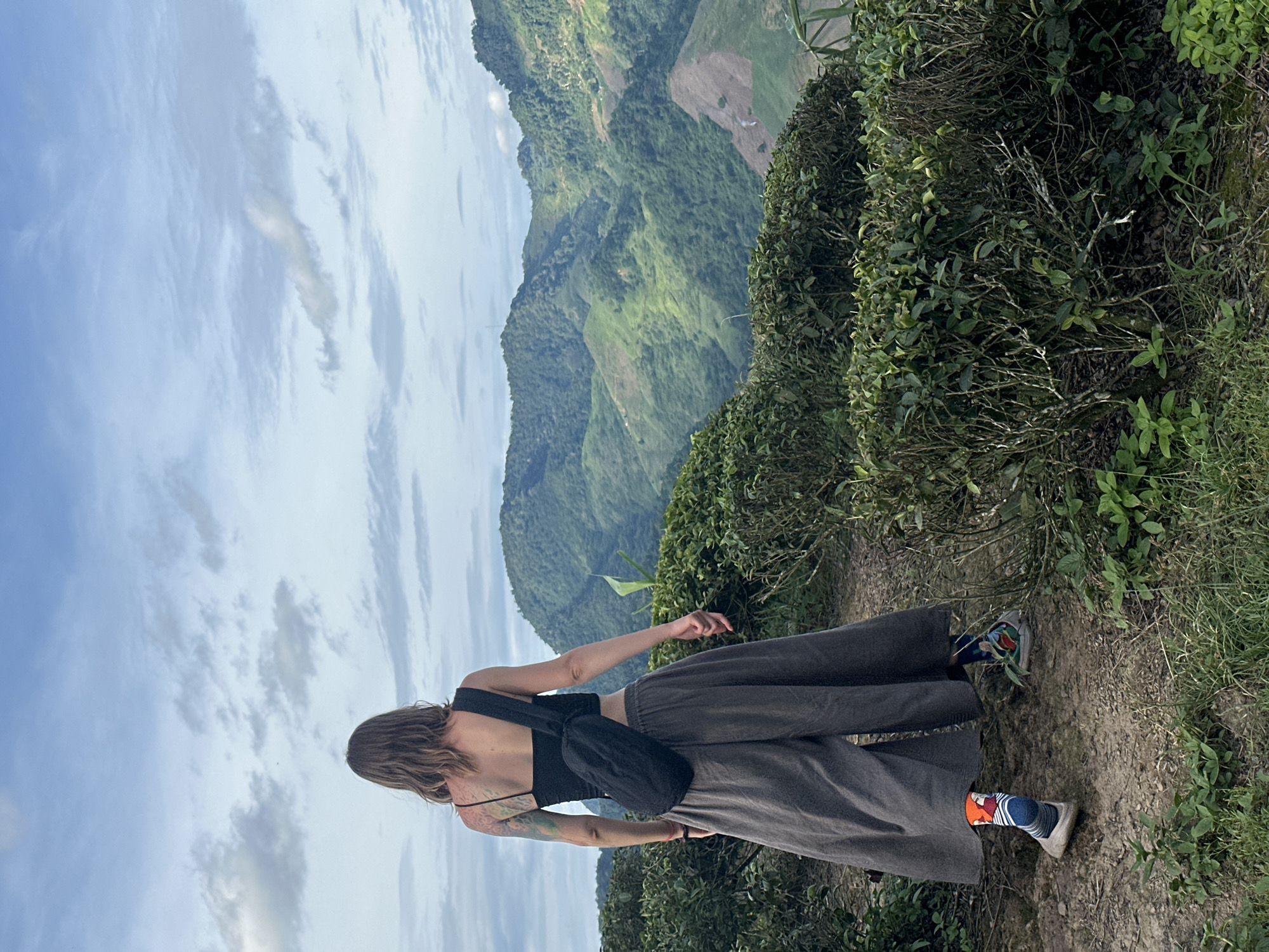 A person wearing a black crop top, gray wide-leg pants, and colorful socks walks on a dirt path through a tea plantation in To Mua, Vietnam. The person has their back to the camera and is carrying a black sling bag across their chest. The background features lush green mountains and a cloudy sky.