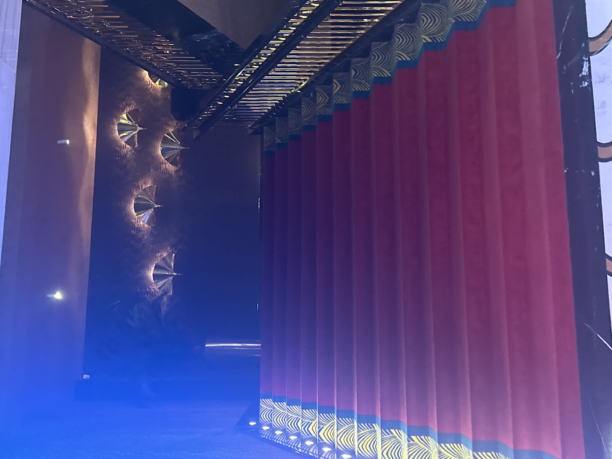 The image shows a grand staircase with a red carpet. The staircase has a black railing with gold accents. The walls are decorated with ornate golden fan-shaped lights. The ceiling is dark and also features decorative lighting. On the left side of the staircase, there is a blue-lit area, possibly a wall or curtain. The overall impression is one of luxury and elegance. The scene is indoors, likely a hotel, theater, or event venue, in Estosadok, Russia. There are no people visible in the image.