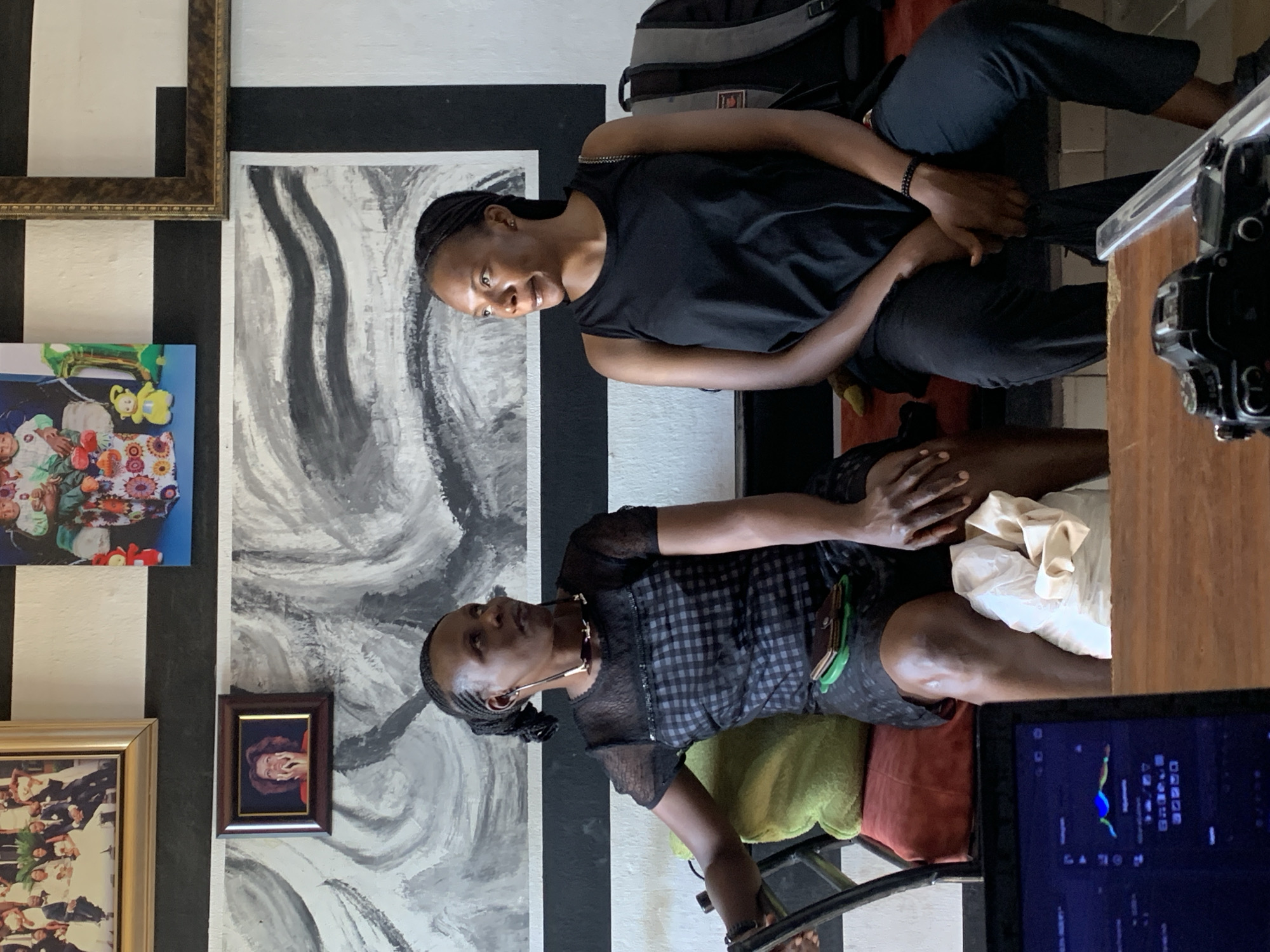 The image captures two women sitting indoors. One woman wears a black tank top and black pants, and has a backpack beside her. The second woman sits on a chair, wearing a dark dress with a subtle pattern and a green accessory at the waist. A laptop rests on a wooden surface in the foreground, displaying data on its screen. Behind the women is a wall decorated with black and white vertical stripes and several framed pictures. An abstract painting hangs on the wall alongside the framed images. The specific location within the city of Jos, Nigeria, is not definitively ascertainable from the visual data alone.