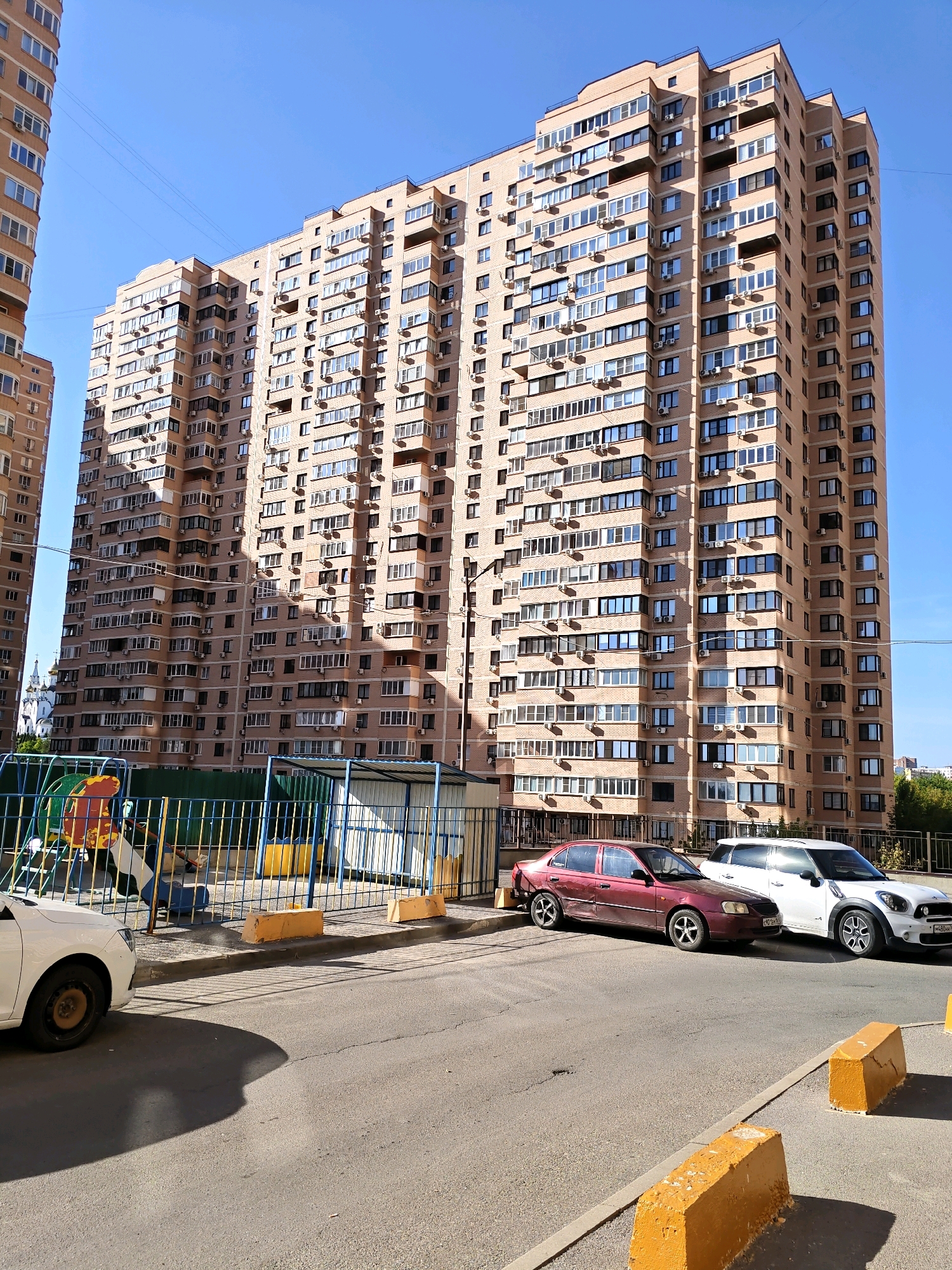 The image captures a residential area in Rostov-on-Don, Russia, under a clear blue sky. A large, multi-story apartment building dominates the scene, its facade clad in light brown brick and adorned with numerous windows and air conditioning units. In the foreground, a playground with colorful structures and a metal fence is visible, suggesting a family-friendly environment. Two cars, a maroon sedan and a white SUV, are parked on the asphalt road in front of the building. The scene is bathed in sunlight, casting shadows on the ground, and the overall impression is one of a typical urban residential setting.