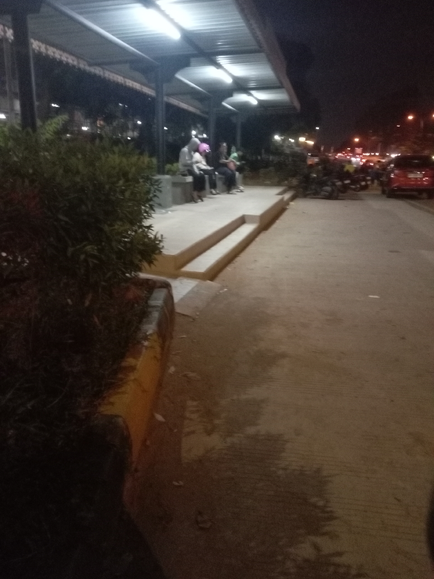 The image depicts a nighttime scene at what appears to be a bus stop or waiting area, likely in Batam, Indonesia, given the context. Under a covered structure illuminated by fluorescent lights, several individuals are seated on a bench. One person is wearing a pink head covering, and others are dressed in casual clothing. Adjacent to the waiting area, there is a patch of dense, dark green shrubbery. A concrete walkway or sidewalk is visible in the foreground, leading towards a road where traffic with illuminated headlights and taillights is present. A red car is parked on the right side of the road. The overall impression is one of a public space during nighttime hours, with people waiting for transportation or services.