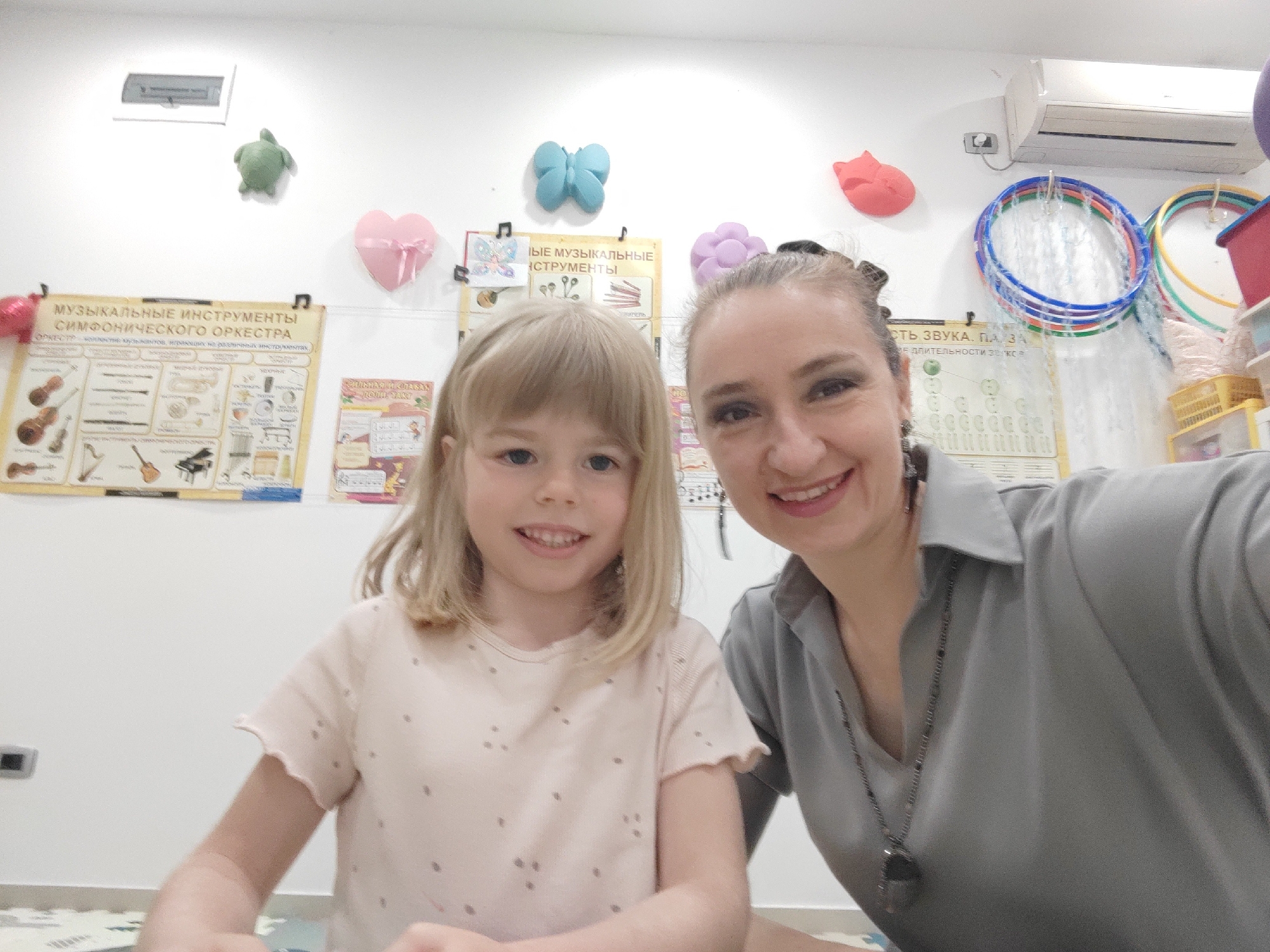 A selfie-style photograph captures an adult woman and a young girl smiling cheerfully indoors, likely in a classroom or children's activity space in Podgorica, Montenegro. The adult, positioned on the right, has dark hair pulled back and is wearing a grey collared shirt with a long necklace. The young girl, on the left, has blonde hair with bangs and is dressed in a light pink t-shirt with small dots. Both are looking directly at the camera with joyful expressions.
The background features a white wall adorned with various decorations and educational materials. Visible wall hangings include a green turtle, a blue butterfly, a pink heart, and a red flower shape. Prominently displayed on the left is a large poster with Cyrillic text, reading "МУЗЫКАЛЬНЫЕ ИНСТРУМЕНТЫ СИМФОНИЧЕСКОГО ОРКЕСТРА" (MUSICAL INSTRUMENTS OF A SYMPHONIC ORCHESTRA), with additional text below. To the right of this, another poster shows "СИЛЬНАЯ И СЛАБАЯ ДОЛИ ЗАКТ" (STRONG AND WEAK BEATS/SHARES...). Further right, another poster mentions "СТЬ ЗВУКА. ПА ЗА МЕ ДЛИТЕЛЬНОСТИ за с" (SOUND. PA... DURATION per s), likely related to music or sound education. An air conditioning unit is mounted on the upper right wall, and several colorful hula hoops hang below it. The floor beneath them appears to be a patterned playmat. The overall atmosphere conveyed is positive and engaging.