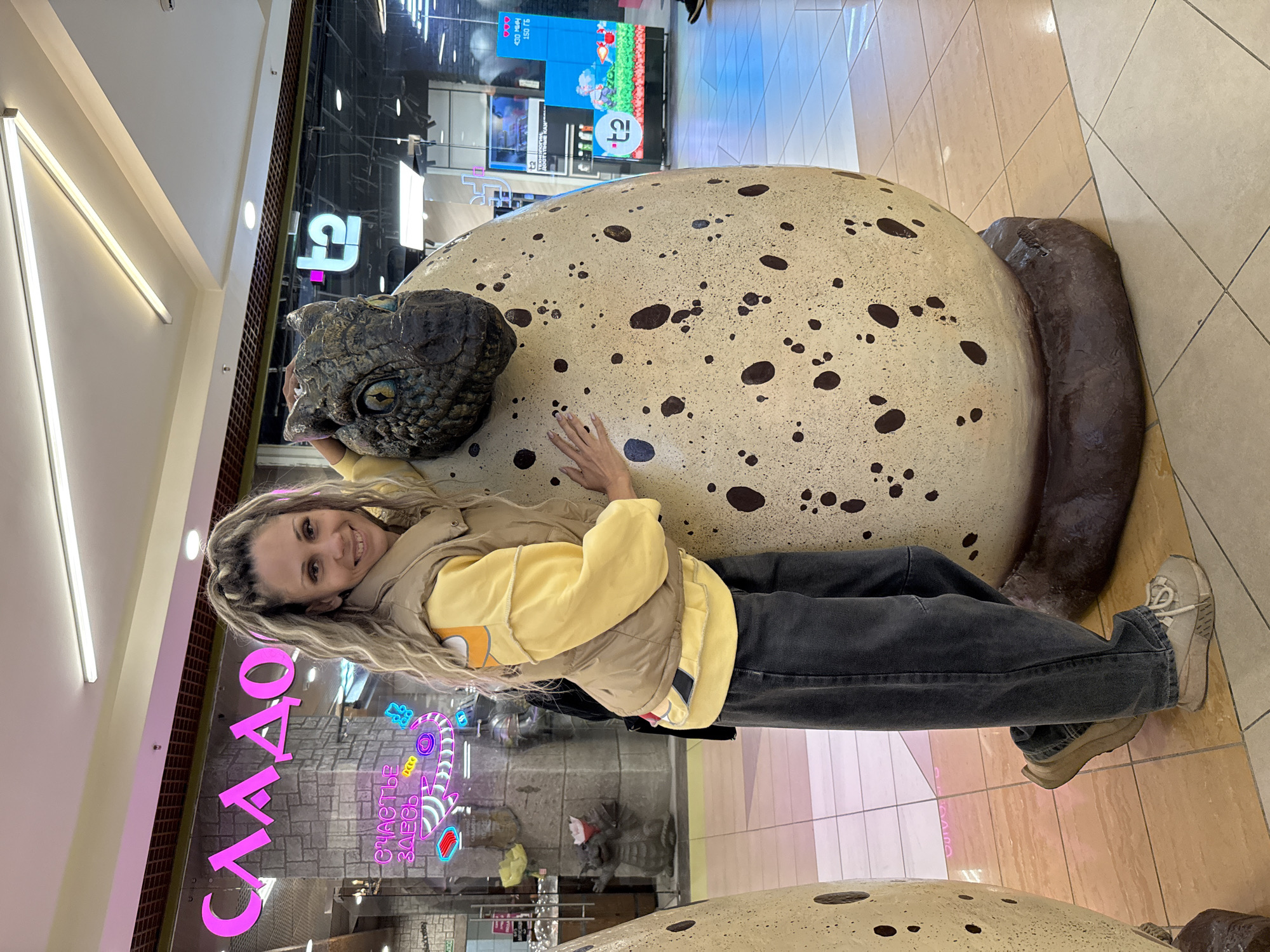 A woman with blonde braided hair is posing next to a large dinosaur egg sculpture with a baby dinosaur head emerging from it. She is wearing a light brown vest over a yellow hoodie and dark gray jeans. Her right hand is resting on the egg, and her left arm is around the dinosaur's neck. The sculpture appears to be in a shopping mall, with shops visible in the background. One shop has neon pink lettering that reads "СЛАДО" which is Russian for "SWEETS". Another shop has a sign with the letters "t2". The floor is tiled in beige and brown squares.