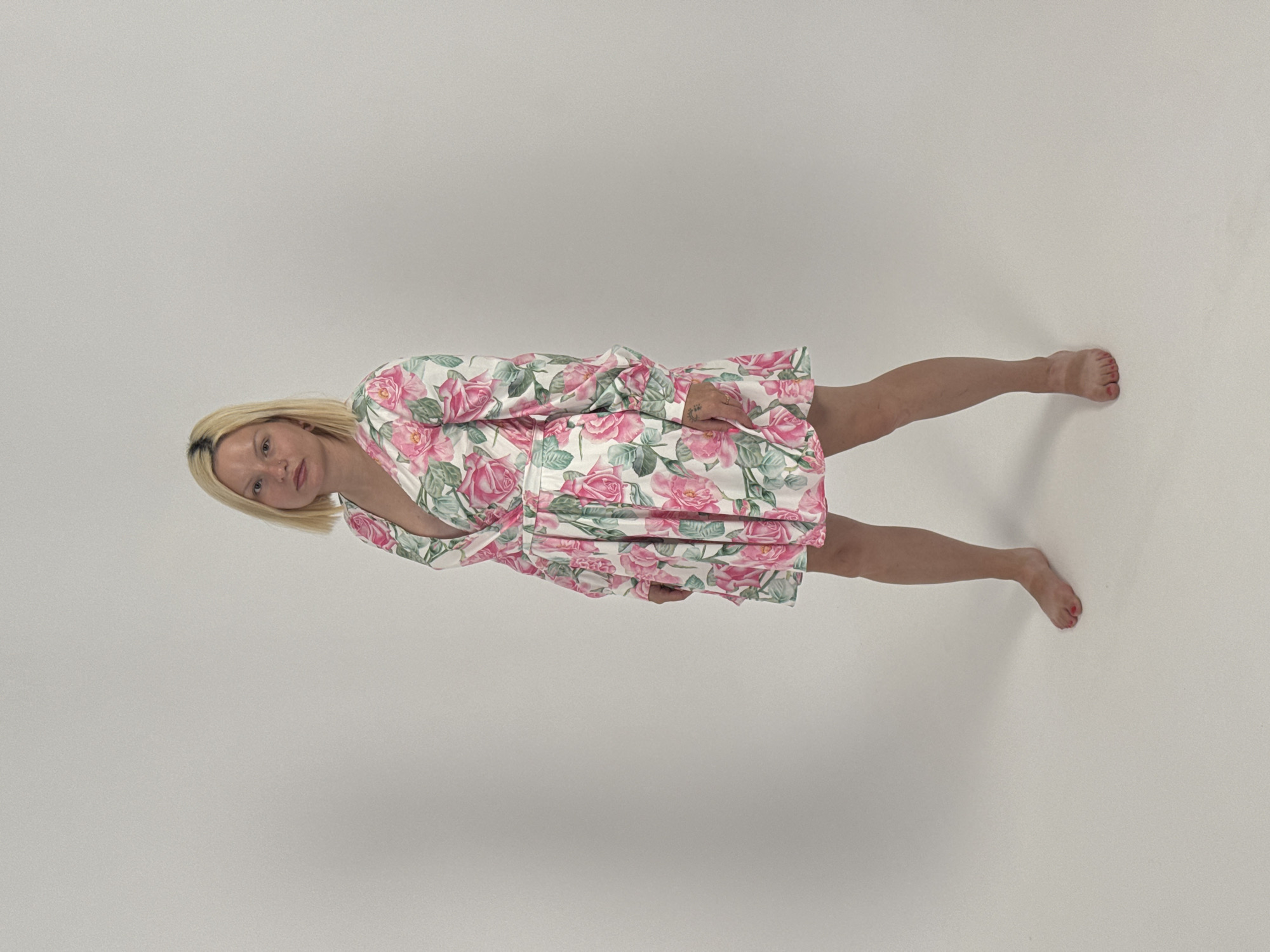An adult individual with blonde, shoulder-length hair stands upright against a plain, light gray background, suggesting an indoor studio setting. The subject is wearing a short, long-sleeved dress adorned with a vibrant pattern of large pink roses and green leaves on a white base. The dress features a V-neckline and appears to be cinched at the waist.
The person is barefoot, and red nail polish is visible on their toes. A small, dark tattoo can be seen on their left wrist or forearm. They face slightly towards the camera, looking directly at the viewer with a neutral and calm expression. Their arms are relaxed by their sides. The lighting is even and bright, highlighting the details of the floral dress. There is no visible text or specific location cues within the image.