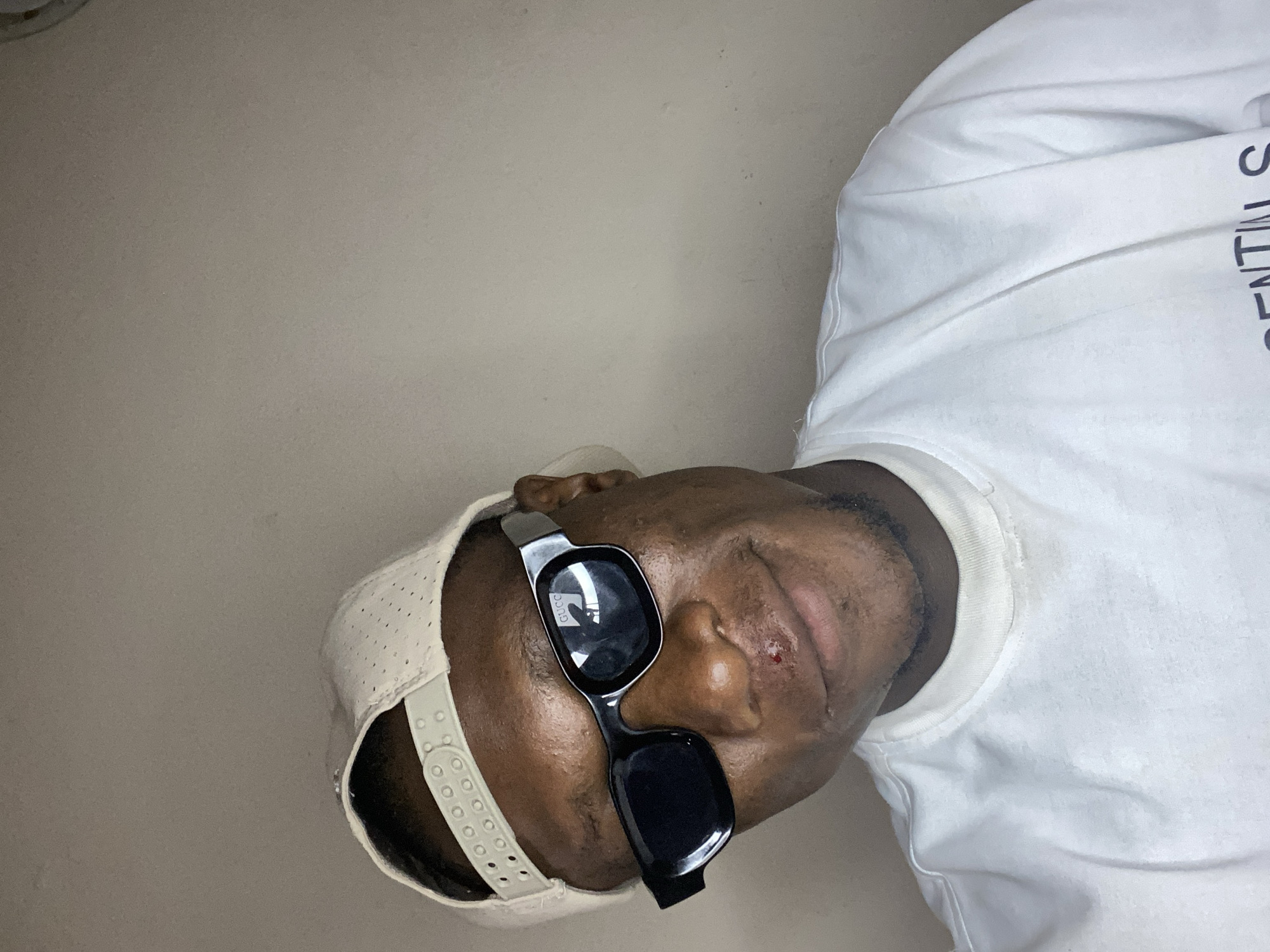 A medium shot depicts a Black man wearing sunglasses and a hat. The man is wearing a white shirt and the sunglasses are black. The background is a plain beige wall. There is no indication the photo was taken in Bukuru, Nigeria.