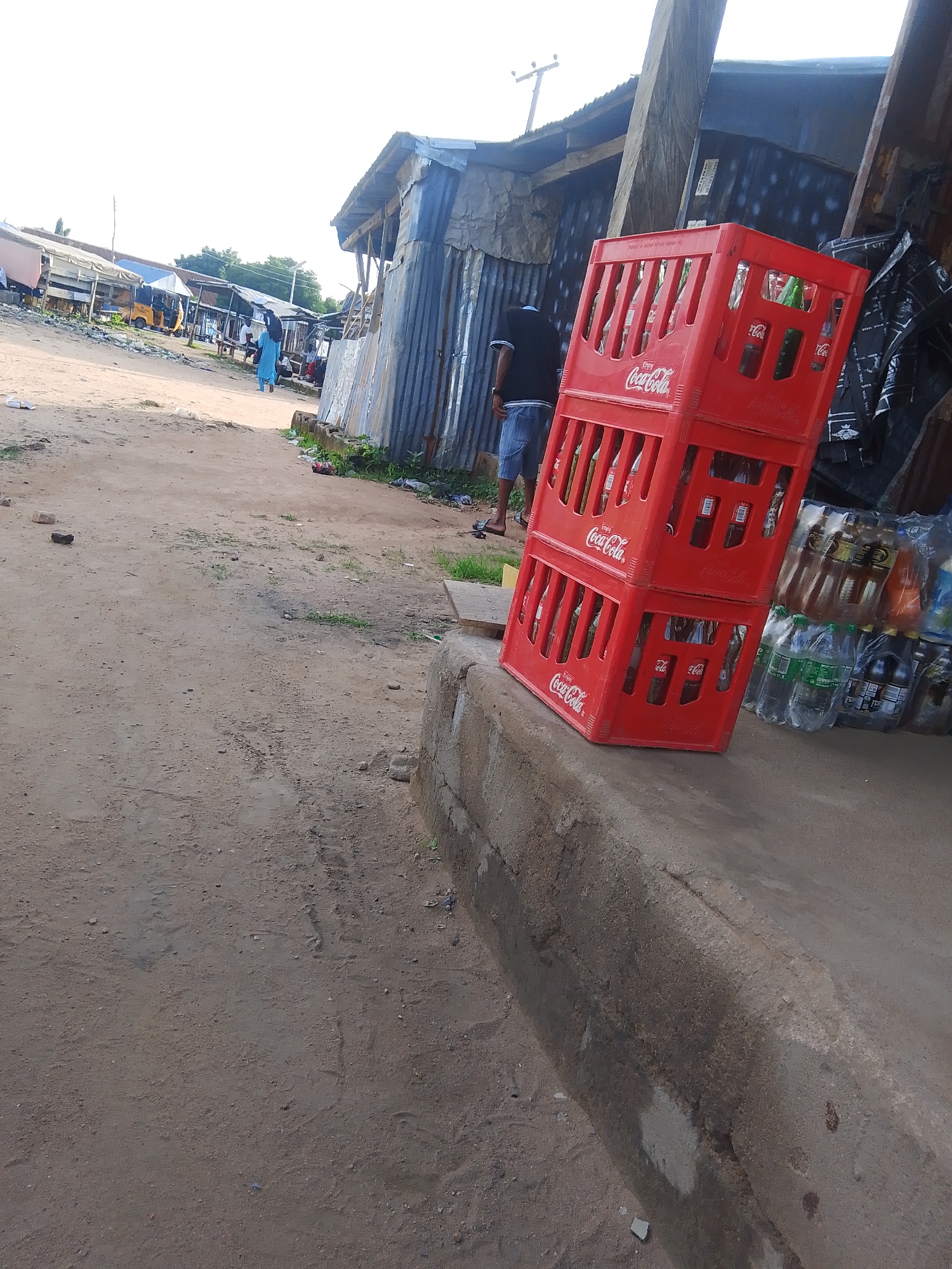 The image depicts an outdoor scene, likely a market or roadside stall in Jalingo, Nigeria. In the foreground, two stacked red Coca-Cola crates filled with bottles are prominently displayed on a concrete ledge. To the right of the crates, a variety of bottled drinks are visible, some in plastic packaging.
In the background, a dirt path leads towards a row of makeshift structures with corrugated metal roofs. A person wearing a blue garment is walking away from the camera on the path, and another person in a dark t-shirt and denim shorts is standing near the crates, facing away. Further in the distance, a yellow motorized tricycle is visible, suggesting transportation common in the area. The scene appears to be set during the day, with bright, overcast conditions. The overall impression is of a bustling, informal commercial area. The text "Coca-Cola" is visible on the red crates.