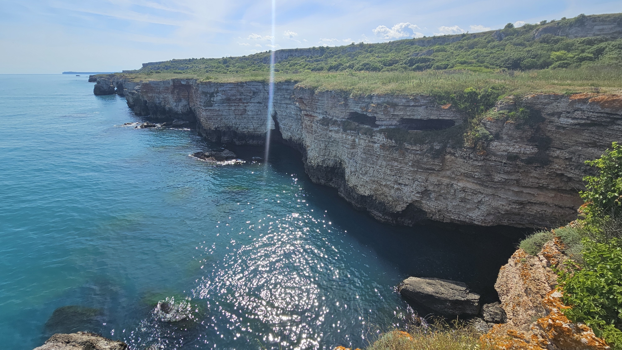 A picturesque view of the Kamen Bryag coastline in Bulgaria unfolds, featuring a series of layered limestone cliffs topped with green vegetation. The teal-colored sea extends to the left, dotted with sun glares. The cliff faces reveal horizontal strata. Erosion has created caves and rocky formations at the base.