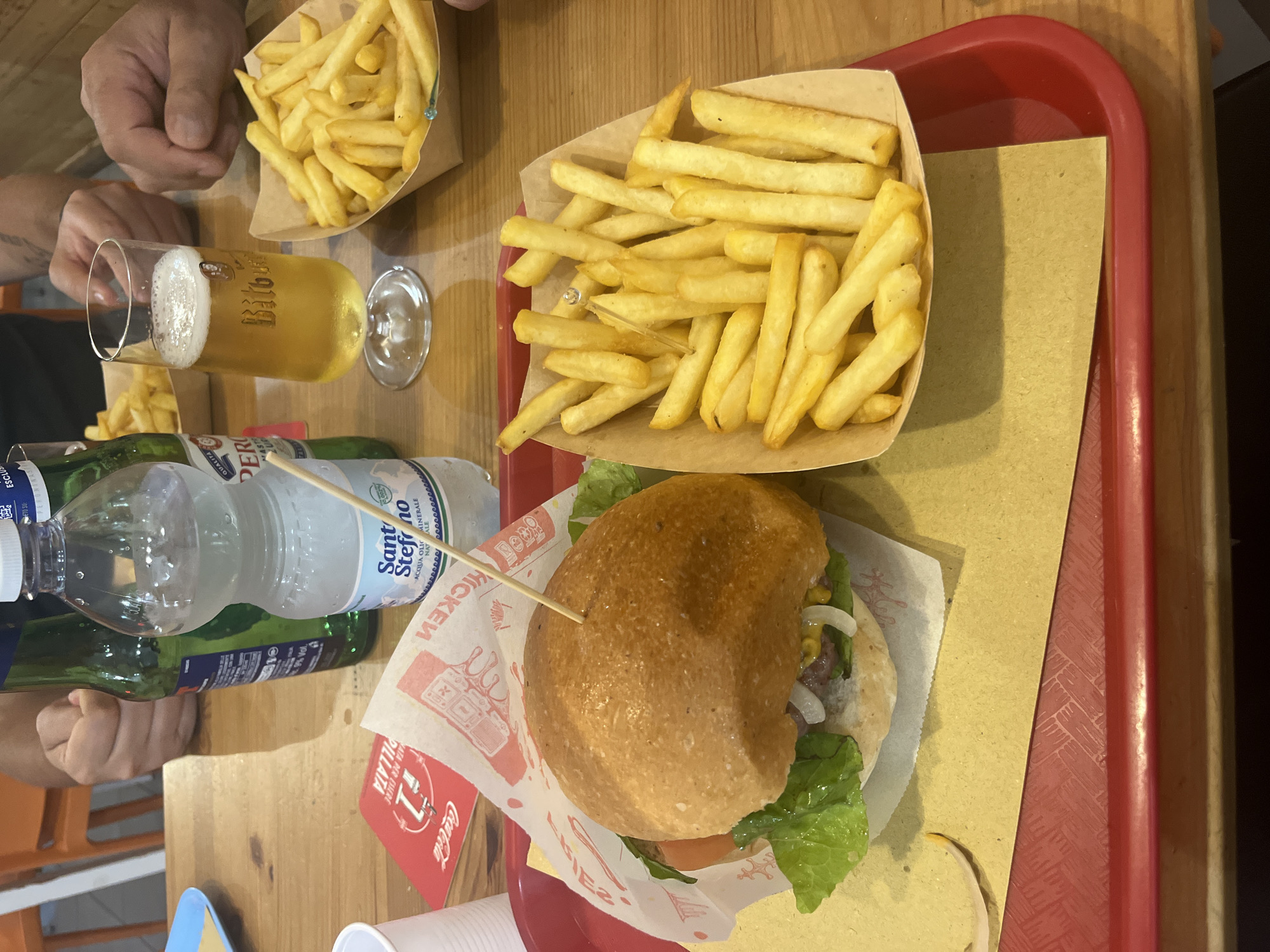 In this medium shot, a wooden table is laden with food and drinks. Two people's hands are visible on the left side of the frame. In the foreground, a hamburger sits on a red tray lined with brown paper. The hamburger has a fluffy bun, green lettuce, and other toppings. Beside it, a bottle of "Santo Stefano" mineral water and a green bottle are placed. A glass of beer, with a prominent "Bitburger" logo, is also on the table, next to a second empty glass. A portion of french fries in a paper container is positioned in the upper portion of the frame, while another portion of fries sits on a separate brown paper wrapper. The scene appears to be a casual meal, possibly at a restaurant or fast-food establishment, given the paper packaging and branding elements like "Coca-Cola" printed on the burger wrapper. The lighting suggests it could be daytime. The overall atmosphere is relaxed and informal. Visible text includes "PILLATA", "HATA PER ESMERE", "1", "Coca-Cola", "9% Vol", "ETO SU", "ESCLUS", "EFONINASE", "QUALITARI", "PER", "MAST", "20", "Santo", "ACQUA OLI MINERALE", "NAT", and "Bitburger". The location context provided indicates Vallo della Lucania, Italy.