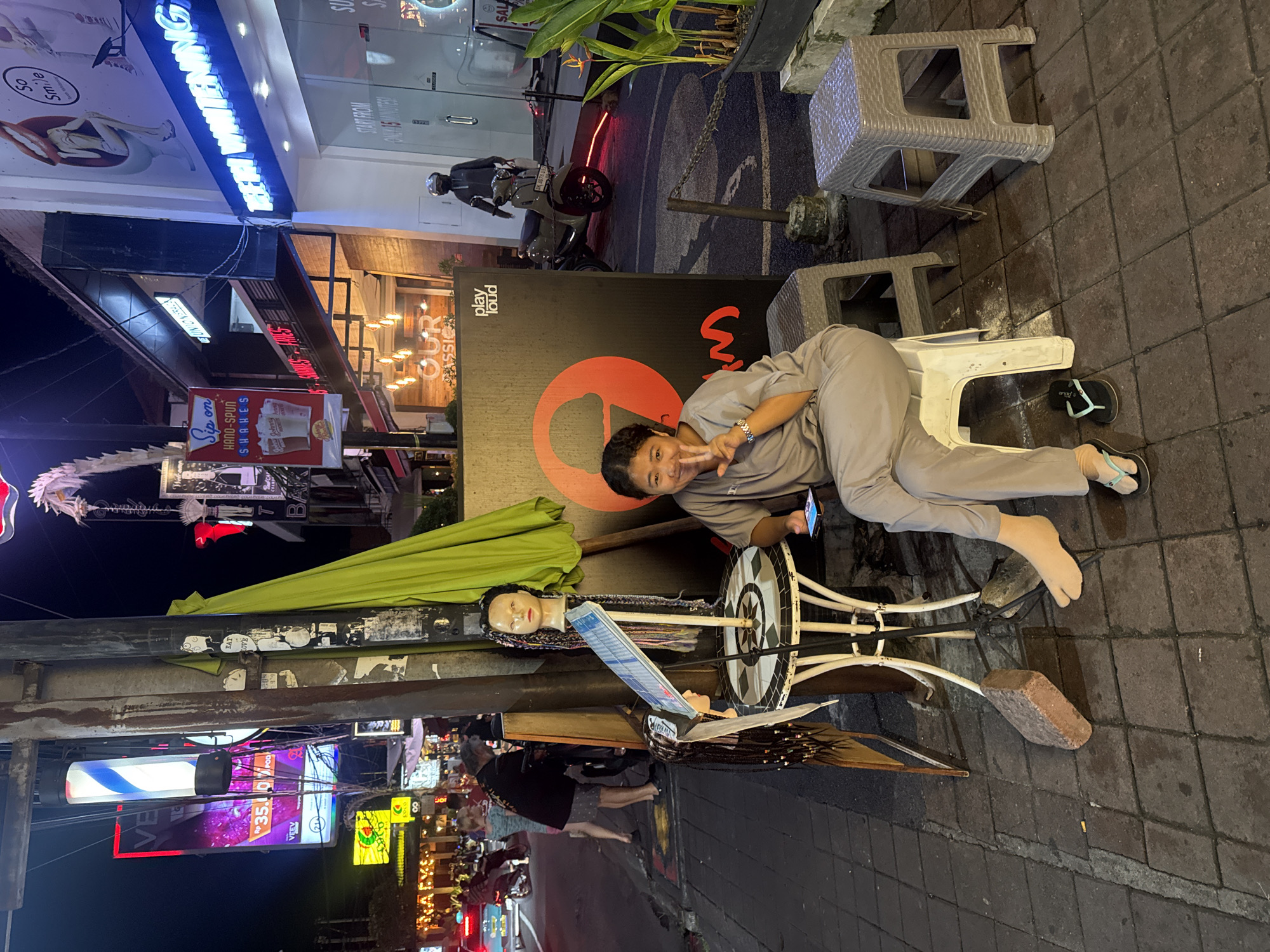 In Kuta, Indonesia, at night, a woman sits on a white plastic stool on a paved sidewalk. She wears a gray jumpsuit and black flip-flops. Her left hand is raised in a peace sign, and she holds a smartphone in her right hand. To her left, a small, ornate metal table displays a mannequin head with braided hair and a sign that reads "Play Loud." Behind her, a large black sign with a red ice cream cone and the text "OUR CLASSIC" is visible. To the right of the woman, a dark gray plastic stool and a potted plant are present. The background shows a street scene with neon signs, including "TEEFI WHITENING" and "Sip on HAND-SPUN SHAKES." A motorcycle is parked near a building.