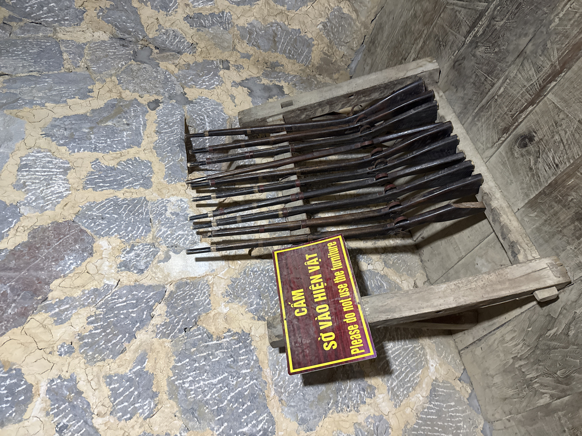 The image shows a collection of antique rifles displayed on a rough wooden rack in what appears to be a museum or historical site. The walls are constructed of stone, and the floor is uneven and textured. A sign in Vietnamese and English reads "Please do not use the furniture," suggesting that the rifles and rack are part of an exhibit. Based on the metadata provided, this image was taken in Sà Phìn Commune, Vietnam.