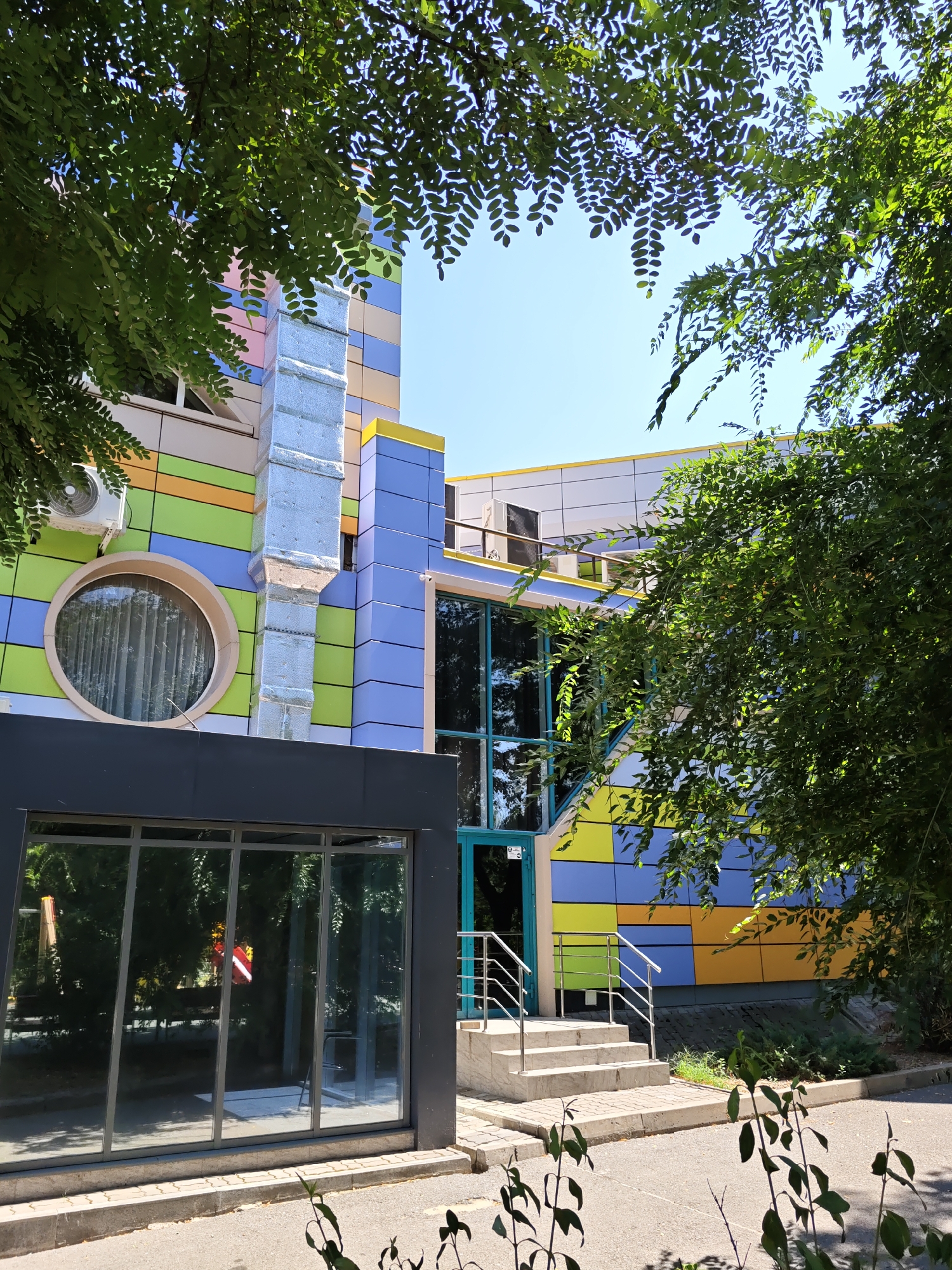 The image shows a modern, colorful building exterior in Rostov-on-Don, Russia. The building features a facade with alternating panels of green, blue, yellow, and light pink. A large, circular window with a beige frame is visible on the left side, to the right of which is an air conditioning unit. A silver-colored duct system runs vertically along the building. To the right, a large glass-fronted section of the building has a teal-framed door and a set of concrete stairs with metal railings leading up to it. The building is partially obscured by dense green foliage from trees in the foreground and on the right side. The ground in front of the building is paved with asphalt and a concrete curb. The sky is clear and blue.