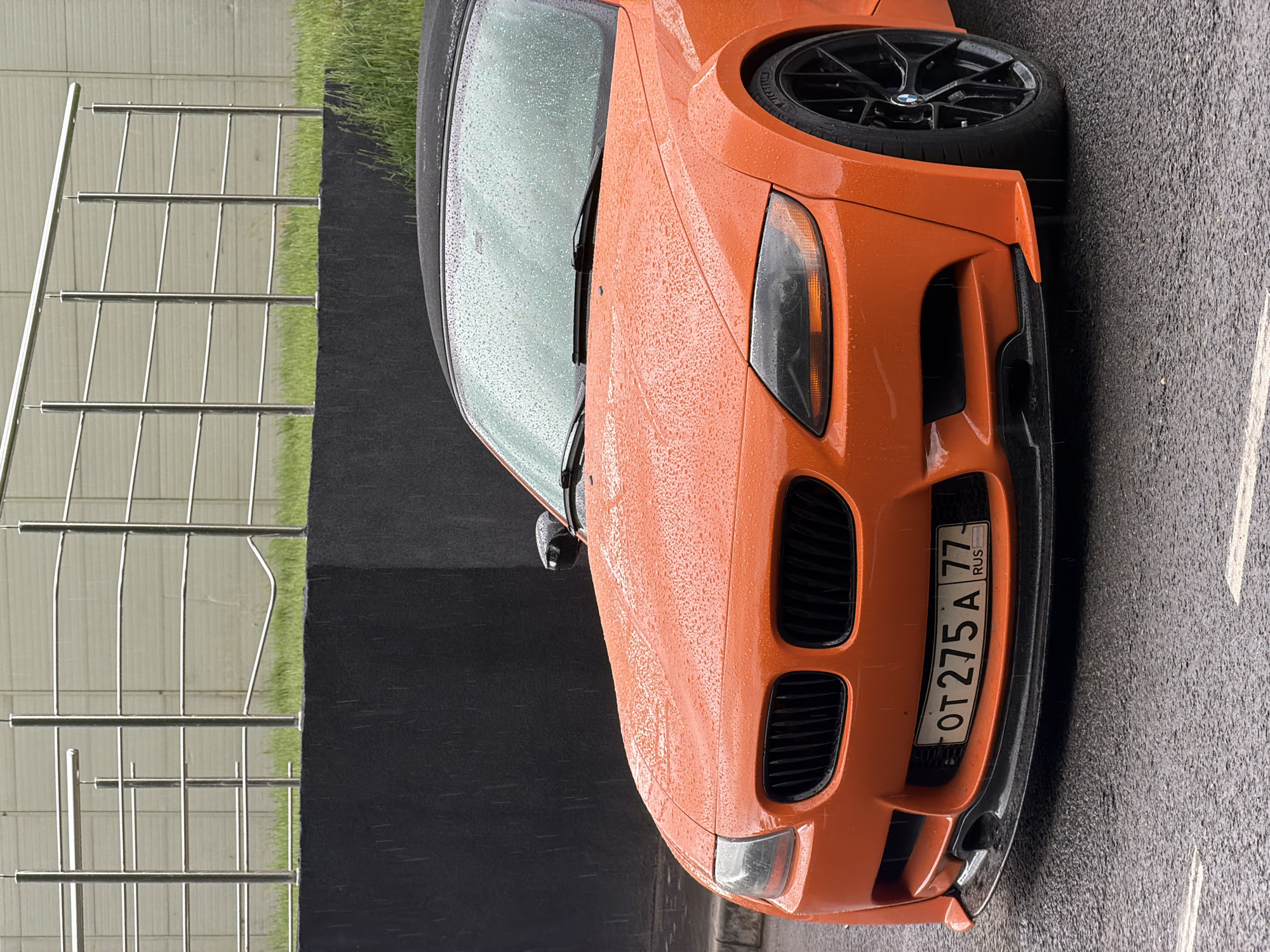 The image shows the front of an orange BMW Z4 car parked on a wet asphalt surface. The car has a Russian license plate that reads "OT275A77 RUS". Raindrops cover the car's surface. A metal fence and a patch of green grass are visible in the background. The location is Moscow, Russia, as indicated by the license plate region code "77". The scene is likely a parking area or street in Moscow.