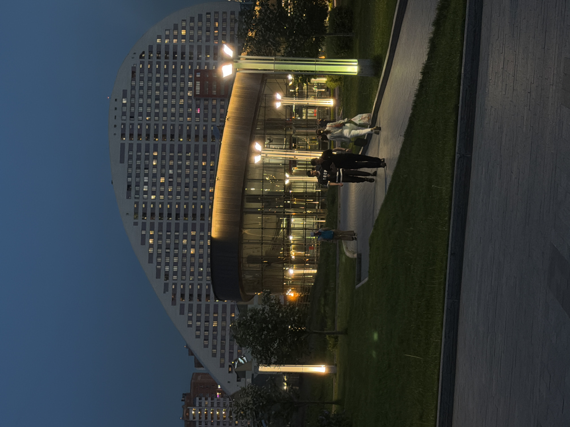 Captured at dusk, the image presents a view within the city of Khimki, Russia. A striking, half-moon-shaped, multi-story building, dotted with numerous lit windows, dominates the scene. Adjacent to this building is a modern structure with curved glass panels, also illuminated.
Several individuals are present. A group of people, possibly a family, are walking along a pathway in front of the modern building. Another person in a blue top is walking in the foreground.
The area is landscaped with patches of green grass, trees, and a paved walkway. Lighting fixtures provide illumination along the path. The sky transitions to a dark blue hue, indicating the approach of night.