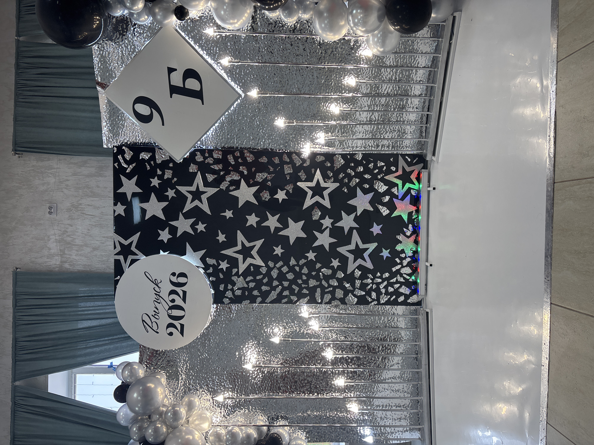 A decorated backdrop for a graduation event is pictured in an indoor setting. The backdrop features a central black panel adorned with silver glitter stars of various sizes. To the left of this is a white circular sign with "Выпуск 2026" (Graduation 2026) written in black script. To the right of the central panel is a white diamond shape with the Cyrillic characters "9 Б" (9 B) in black. Both the central black panel and the white diamond are flanked by sections of silver, crinkled reflective material. A cluster of silver and black balloons is visible on the far left and right edges of the backdrop. The backdrop is illuminated by vertical silver stands with small, glowing lights. A white, reflective floor is present in the foreground. In the background, teal curtains are drawn across a window, and a textured grey wall with a light switch is visible. The scene is likely a school gymnasium or event hall in the city of Chaikovsky, Russia, set up for a graduation ceremony.