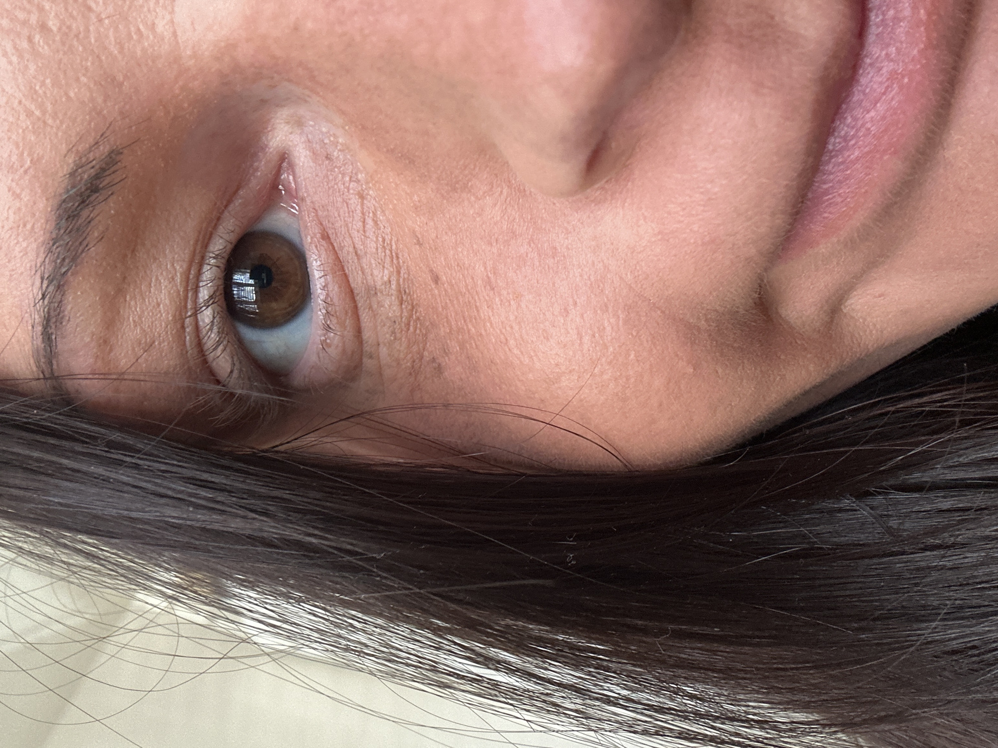 This close-up portrait focuses intensely on a person's face, primarily showcasing their right eye, nose, and part of their lips, with dark hair framing the lower section of the image. The subject's head appears tilted, suggesting they might be lying down or in a reclined position.
The eye features a rich brown iris with a distinct black pupil. Reflections within the pupil indicate an indoor light source, possibly a window or a lamp. Dark, prominent eyelashes frame the eye, and a portion of a well-defined dark eyebrow is visible above it. The skin around the eye, nose, and forehead displays natural texture with visible pores and fine lines, and has a warm, peachy-tan complexion.
To the right, a segment of the nose bridge and nostril is visible, along with a part of the upper lip which has a pinkish hue, suggesting either natural color or light lip product, and some visible texture.
A mass of straight, dark brown or black hair sweeps across the bottom of the frame, resting on what appears to be a light-colored surface, such as bedding or fabric. Individual strands of hair are clearly discernible.
The image captures an intimate and detailed perspective, bathed in soft, even lighting, conveying a sense of calm or introspection. There is no visible text.