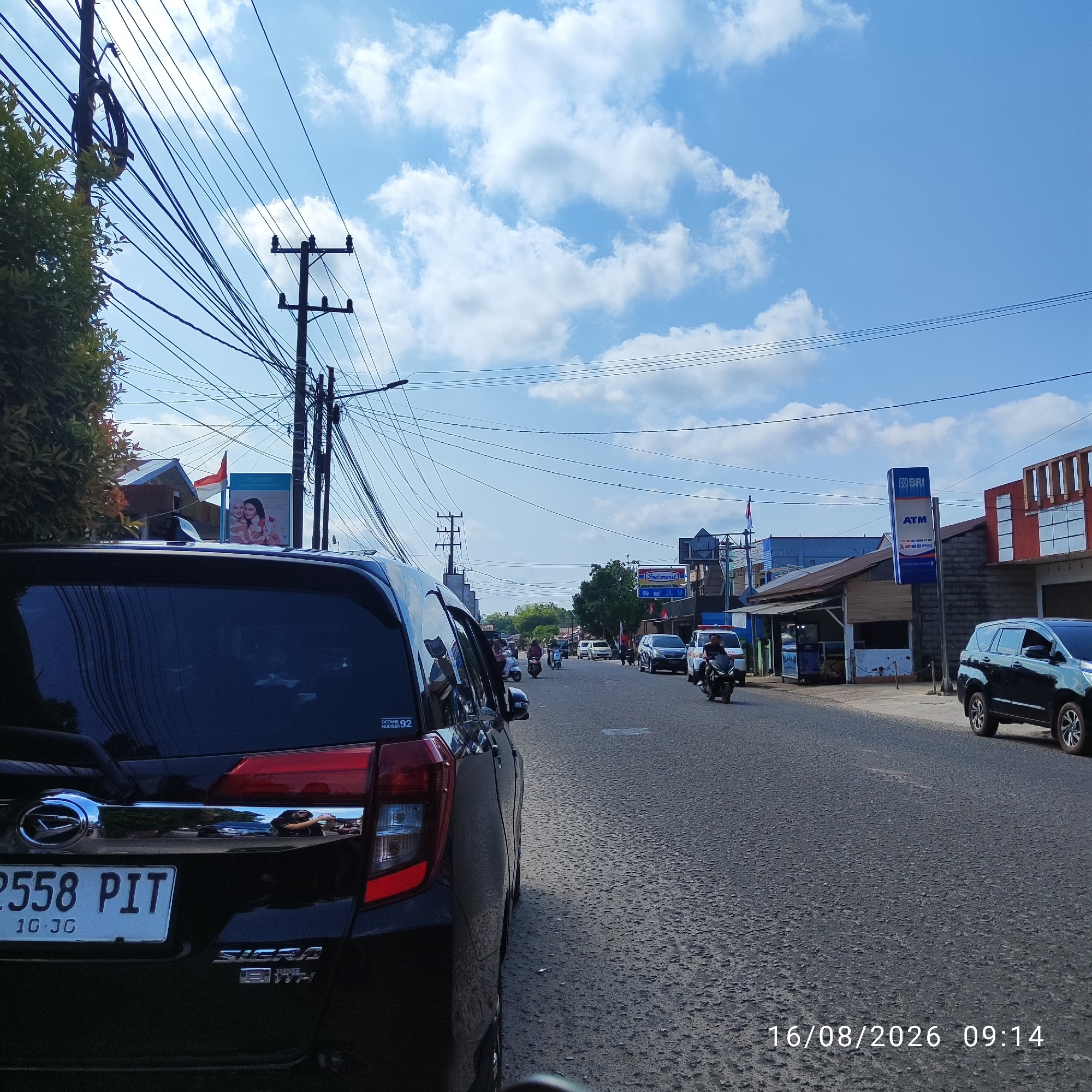The image depicts a street scene in Pembaharuan, Indonesia, on August 16, 2026, at 09:14. The perspective is from within a dark-colored vehicle, looking forward along a road with a paved surface. Several vehicles, including cars, motorcycles, and a van, are present on the road, indicating moderate traffic. A black SUV is parked on the right side of the road, and another black vehicle, partially visible, is in the foreground on the left.
The environment includes buildings lining the street, with one notable building displaying a "BRI ATM" sign. Utility poles with a dense network of overhead wires are visible on both sides of the street, characteristic of urban infrastructure. Lush green trees and foliage are present on the left side of the image. The sky is predominantly blue with scattered white clouds.
A close-up of the rear of the black vehicle in the foreground reveals a license plate reading "2558 PIT" and "10 JG," along with "SIGRA" and "1.2" badging. A sticker on the rear window reads "OCTANE NUMBER 92". In the background, on the left, a billboard with "Wardah" branding is visible, featuring images of two women and floral elements, alongside an Indonesian flag.