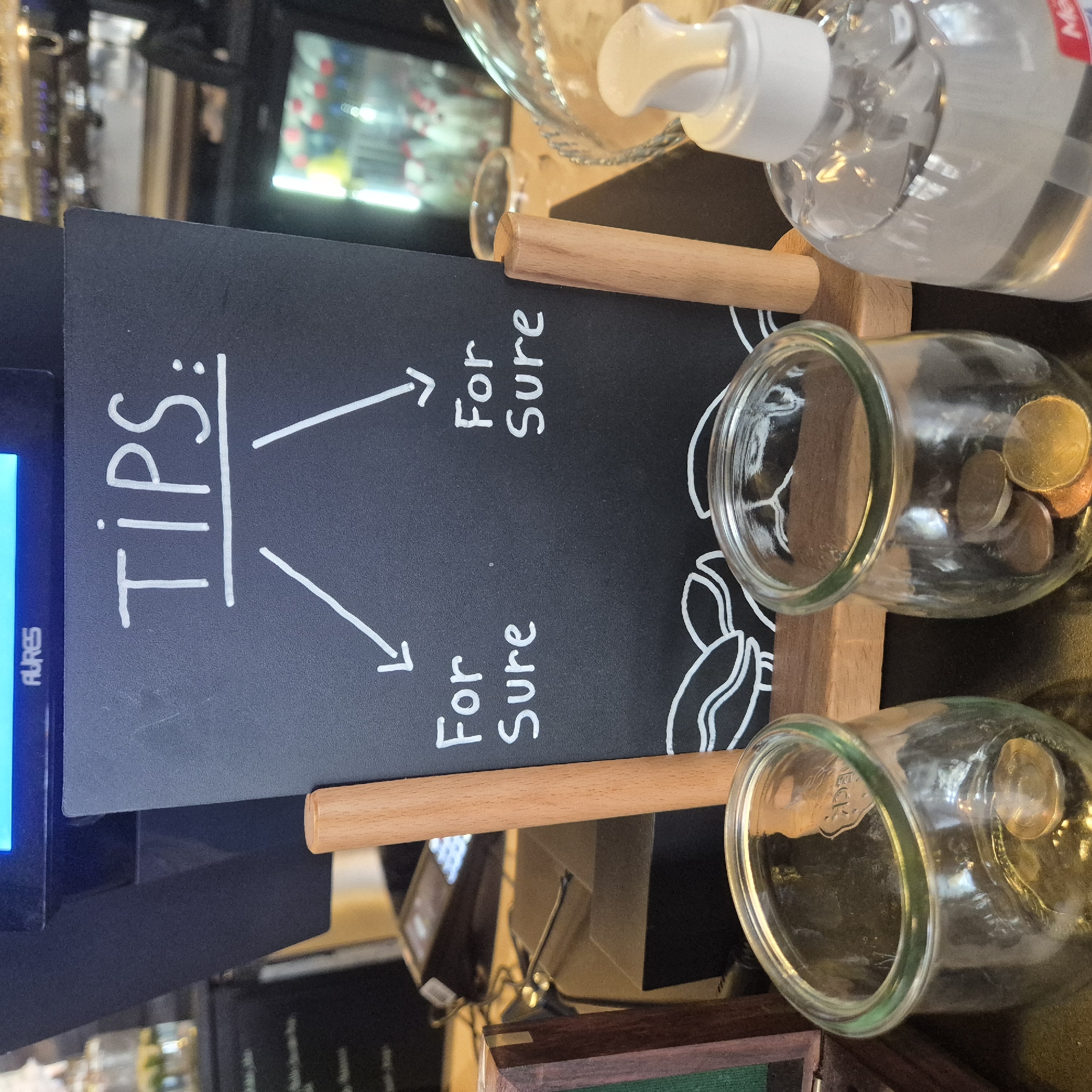The image depicts a tip jar setup at a business, likely in Strasbourg, France. A black chalkboard sign reads "TIPS:" with two arrows pointing downwards, each labeled "For Sure". Below the sign are two glass jars with some coins inside. The jars are supported by a wooden stand with horizontal wooden dowels. A bottle of hand sanitizer is positioned nearby. The surrounding environment suggests a cafe or restaurant setting.