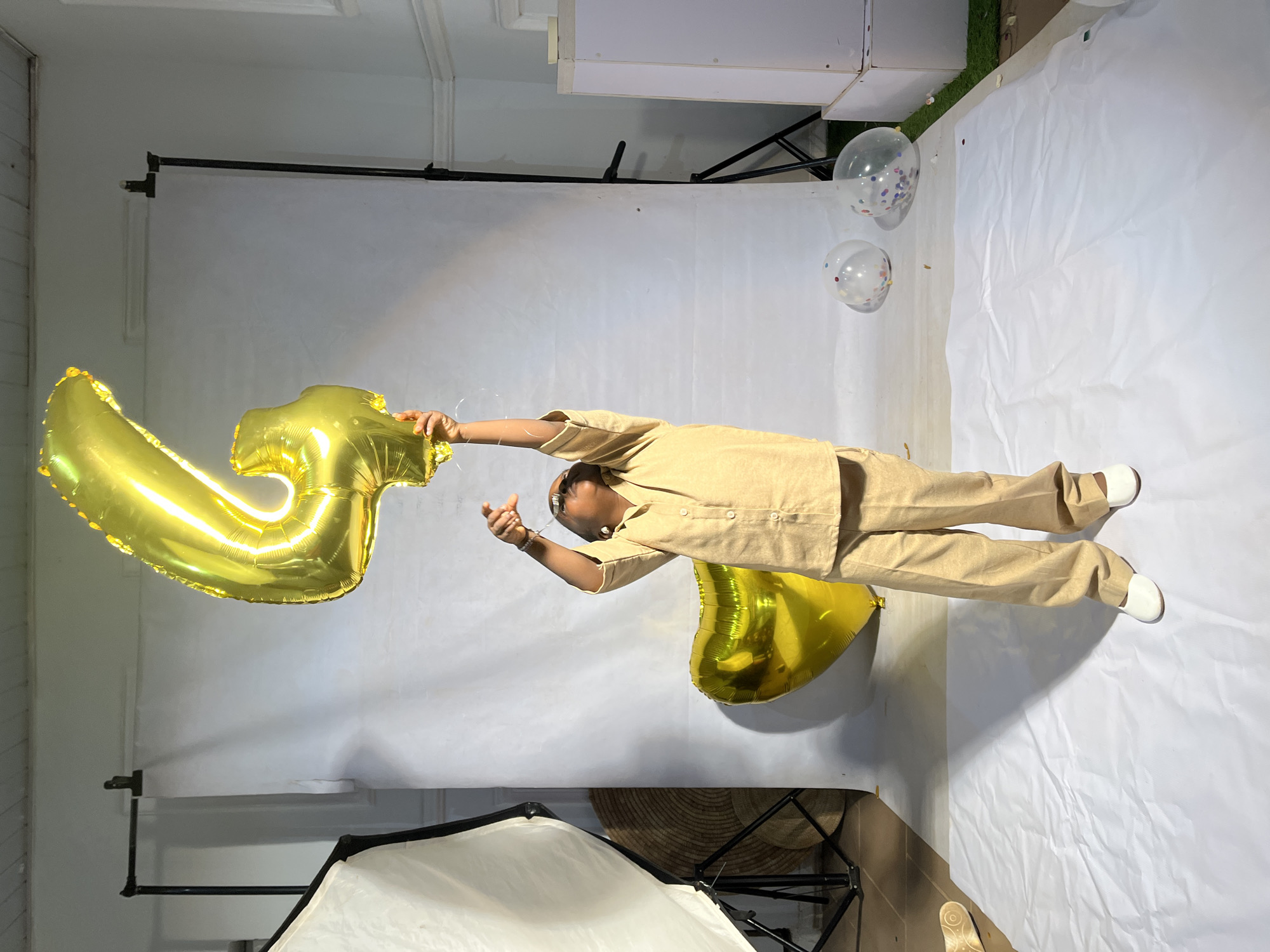 In a studio setting, likely in Jos, Nigeria, a young child stands with arms raised, holding a large, gold, Mylar balloon shaped like the number "4". The child wears a tan, collared shirt and matching loose-fitting pants, completed by white shoes. Behind the child, a large gold heart-shaped balloon rests on the white paper-covered floor. The background consists of a plain white backdrop, with photographic lighting equipment visible on the left. Two clear balloons, containing colorful confetti, are positioned on the floor to the right of the child. The scene suggests a birthday or celebration photoshoot.