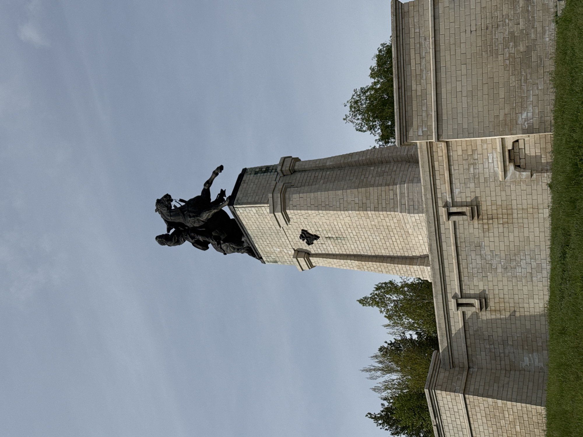 The image depicts the Soldiers' and Sailors' Memorial Bridge in Harrisburg, Pennsylvania. The bridge features a tall, brick structure topped with a bronze equestrian statue. The sky is visible in the background. A small amount of green grass and foliage are also present.