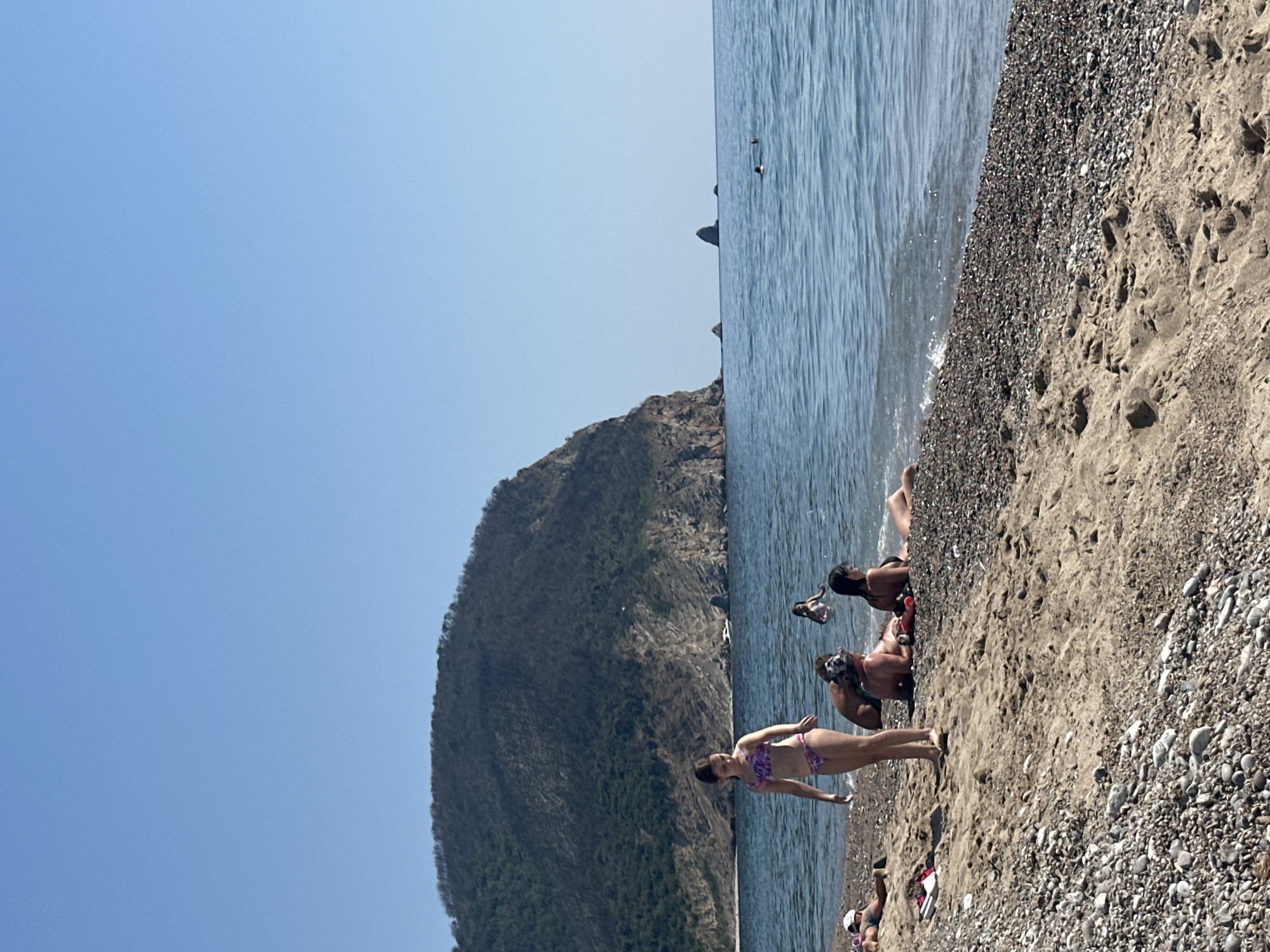 The image depicts a sunny day at a beach, likely in Đurovići, Montenegro. The scene is divided horizontally: the top portion shows a clear, bright blue sky, transitioning to the shimmering blue water of the Adriatic Sea. The bottom portion of the image is dominated by a sandy and pebbly beach, with a large, tree-covered mountain rising dramatically on the left side, bordering the sea.
Several people are visible enjoying the beach. In the foreground, a woman in a patterned bikini stands on the sand, facing away from the camera. Further back, a group of people are relaxing and interacting on the shore. Some are seated, while others appear to be in the water. One person is in the process of diving or jumping into the water, with their body partially submerged. The presence of people in swimwear and the overall relaxed atmosphere suggest a warm, pleasant day suitable for swimming and sunbathing.
The water appears calm with gentle waves lapping at the shore, and the sunlight creates a bright, summery ambiance. The texture of the beach, a mix of fine sand and small pebbles, is clearly visible. The mountain's dense vegetation suggests a lush, natural landscape. No visible text is present in the image. The overall impression is one of a leisurely day at a beautiful coastal location.