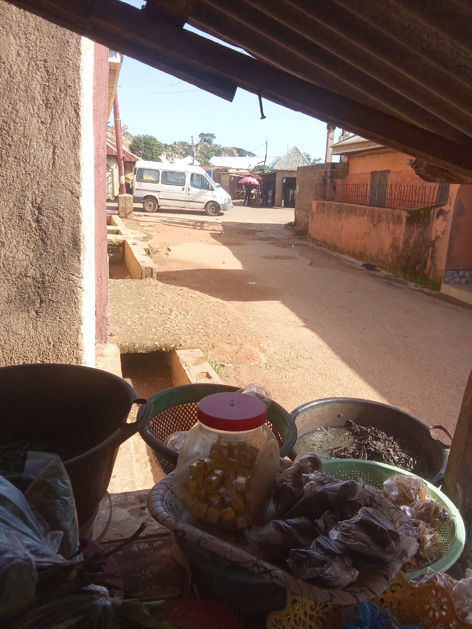 In an outdoor market stall, under a corrugated metal roof, various goods are displayed. In the foreground, a large black plastic bucket sits next to a green plastic basket filled with wrapped items. A clear glass jar containing yellow cube-shaped objects with a red lid is also present. In another green basket, dried leafy greens are submerged in murky liquid within a large dark bowl. The stall opens onto a street with a white van parked on the right side of the road. Buildings with orange walls and various architectural styles line the street, suggesting an urban or semi-urban setting. The ground is paved with reddish-brown dirt and gravel. The sky is clear and blue. The scene appears to be from a street-side vendor in a possibly warm climate.
