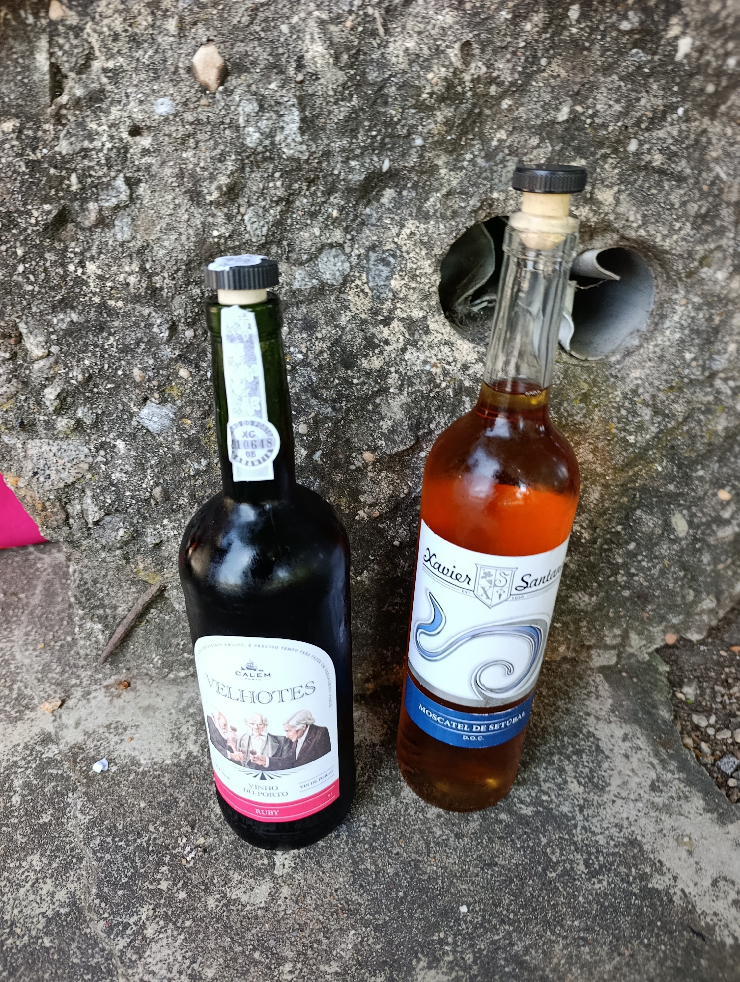 Two bottles of wine are on a rough concrete surface. The bottle on the left is dark green and has a label with a blue and white design featuring three men. The label also reads "CALEM VELHOTE'S VINHO DO PORTO RUBY". A small sticker on the neck of the bottle has the text "XC 10648 05". The bottle on the right is clear glass and contains a reddish-brown liquid. Its label is white and blue and says "Xavier Santan", "EST 1920", and "MOSCATEL DE SETUBAL D.O.C.". The bottles are positioned near a circular opening in the concrete, which appears to be a drain or ventilation hole. The concrete surface is textured and appears to be outdoors. The image is taken from a high angle, looking down at the bottles. The location is identified as Trofa, Portugal.