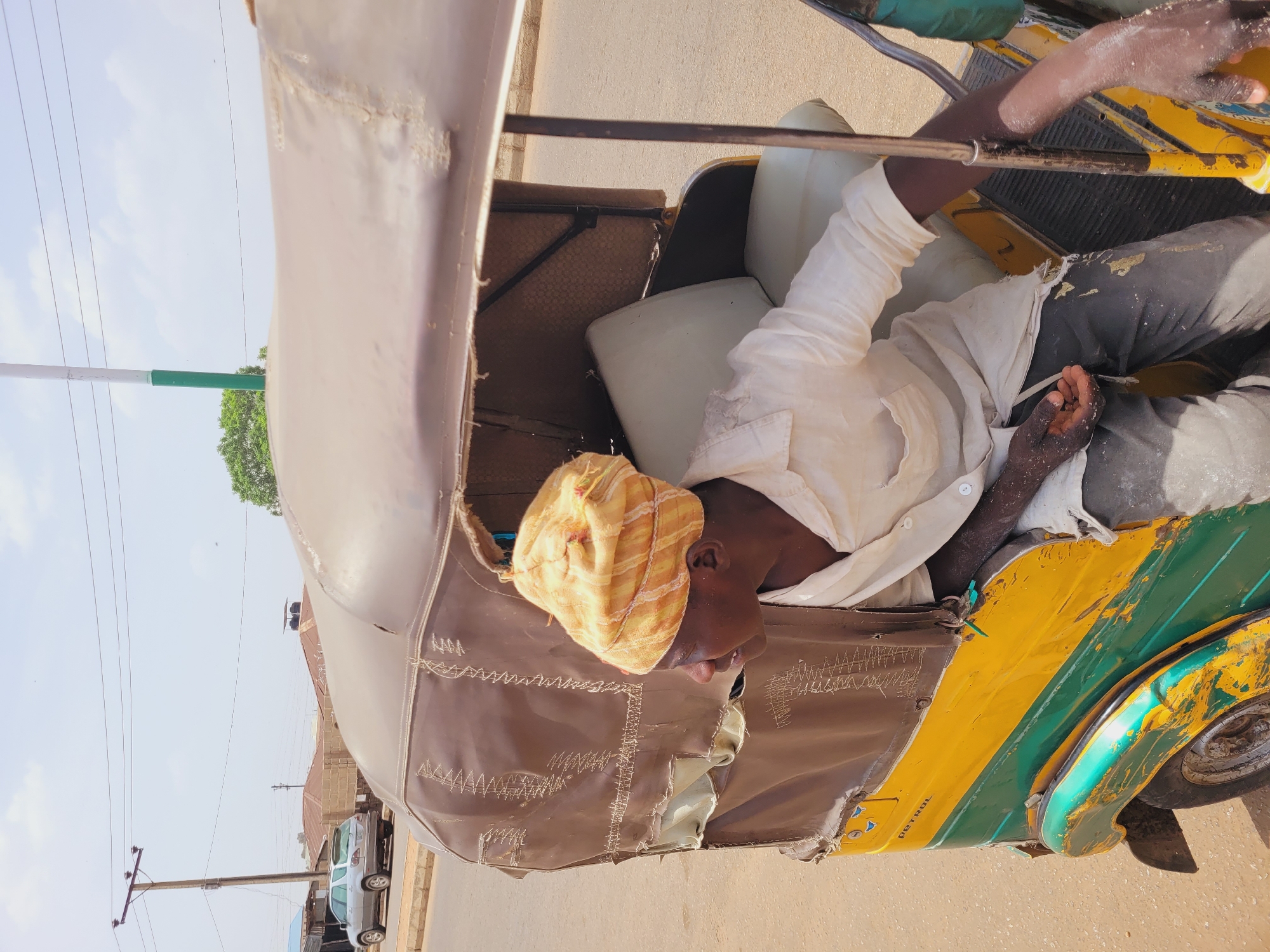 An adult male, likely in his later years, is seated inside a yellow and green three-wheeled vehicle, identified as a tricycle, or "keke," a common mode of transport in Nigeria. The man is wearing a light-colored, long-sleeved shirt and dark pants. He has a patterned yellow and orange cap on his head. The keke has a tan-colored canvas cover with what appears to be stitching detail. The photograph, taken during daylight hours, shows the vehicle on a paved road. Other buildings, vehicles, and utility poles can be seen in the background, suggesting an urban environment. The image was taken in Bukuru, Nigeria.
