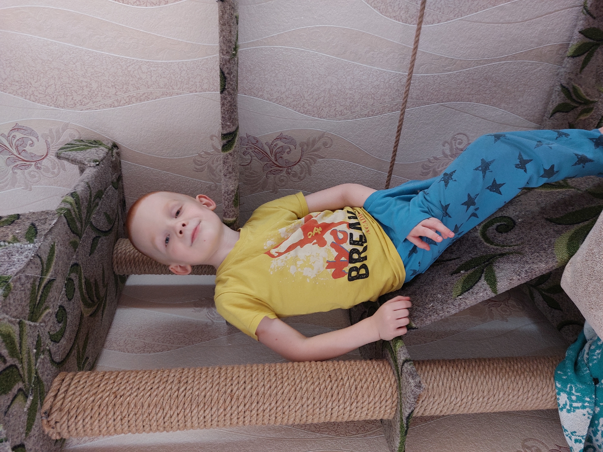 A child with red hair is lying on a multi-tiered structure. The child is wearing a yellow t-shirt with a print and blue pants with black stars. The structure has carpeted steps and posts wrapped in rope. The background features a beige patterned wall. This photograph may have been taken in Большой Букор, Russia, as it was previously mentioned.