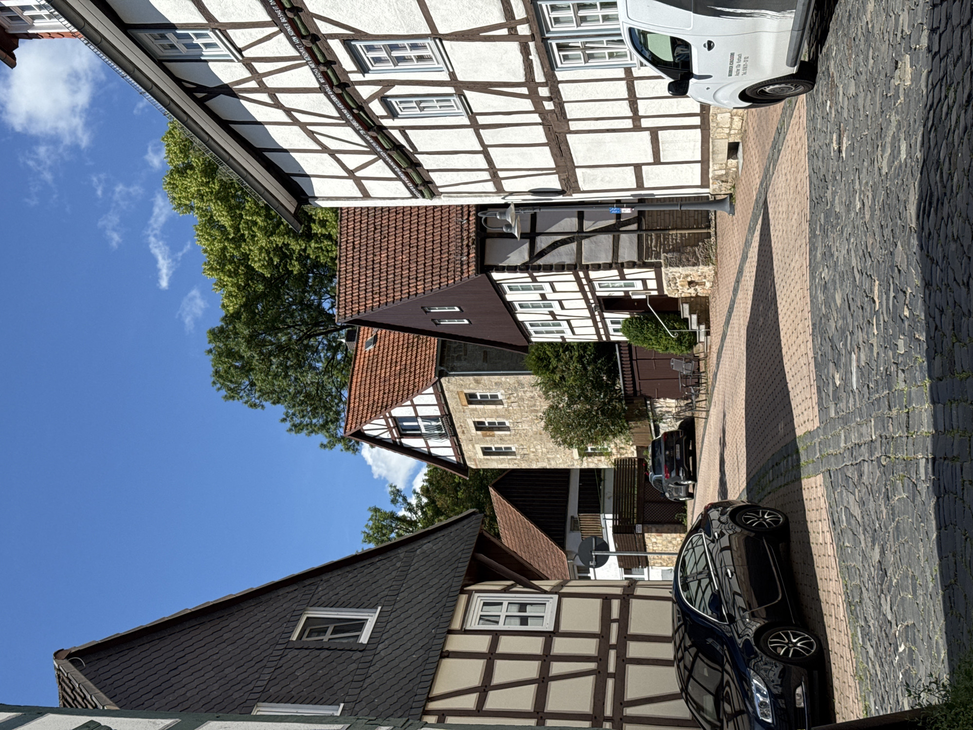 In an exterior, eye-level, medium shot, a cobblestone street curves through a historic European village on a clear, sunny day. The street is lined with traditional timber-framed houses with white stucco infill and dark brown timber beams. On the left, a dark sedan is parked on the street. Across the street, a white van is parked near the curb. In the background, trees and a blue sky with scattered clouds are visible. The architecture suggests a historical European setting, possibly Germany, given the timber-framed construction style.