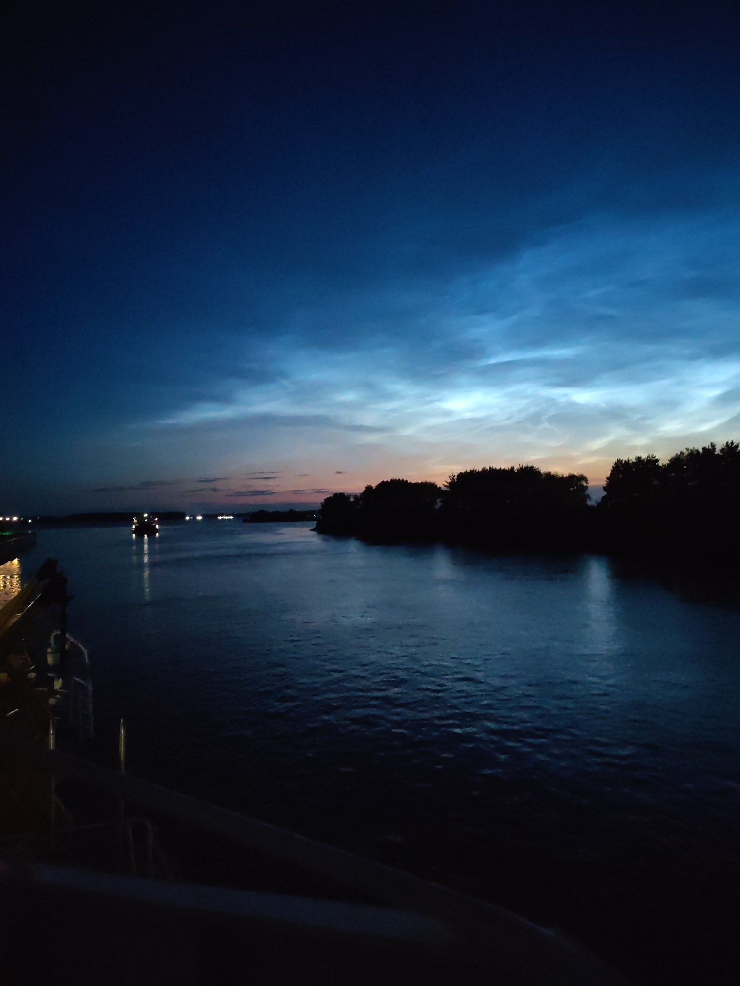 The image depicts a wide river at dusk, with a dark blue sky overhead featuring wispy, luminous clouds. Silhouetted trees form a dark line along the right bank of the river. In the distance, on the water, several small boats with lights are visible. The foreground shows a portion of a vessel, likely a ferry or cargo ship, with railings and structures in dark silhouette. The water is calm, reflecting the dim light of the sky and the boat lights. The overall atmosphere is tranquil and serene, characteristic of twilight on a body of water. The scene is consistent with a location along a river, such as the Lena River near the settlement of Bol'shie Mkhi, Russia, where such vistas are common.