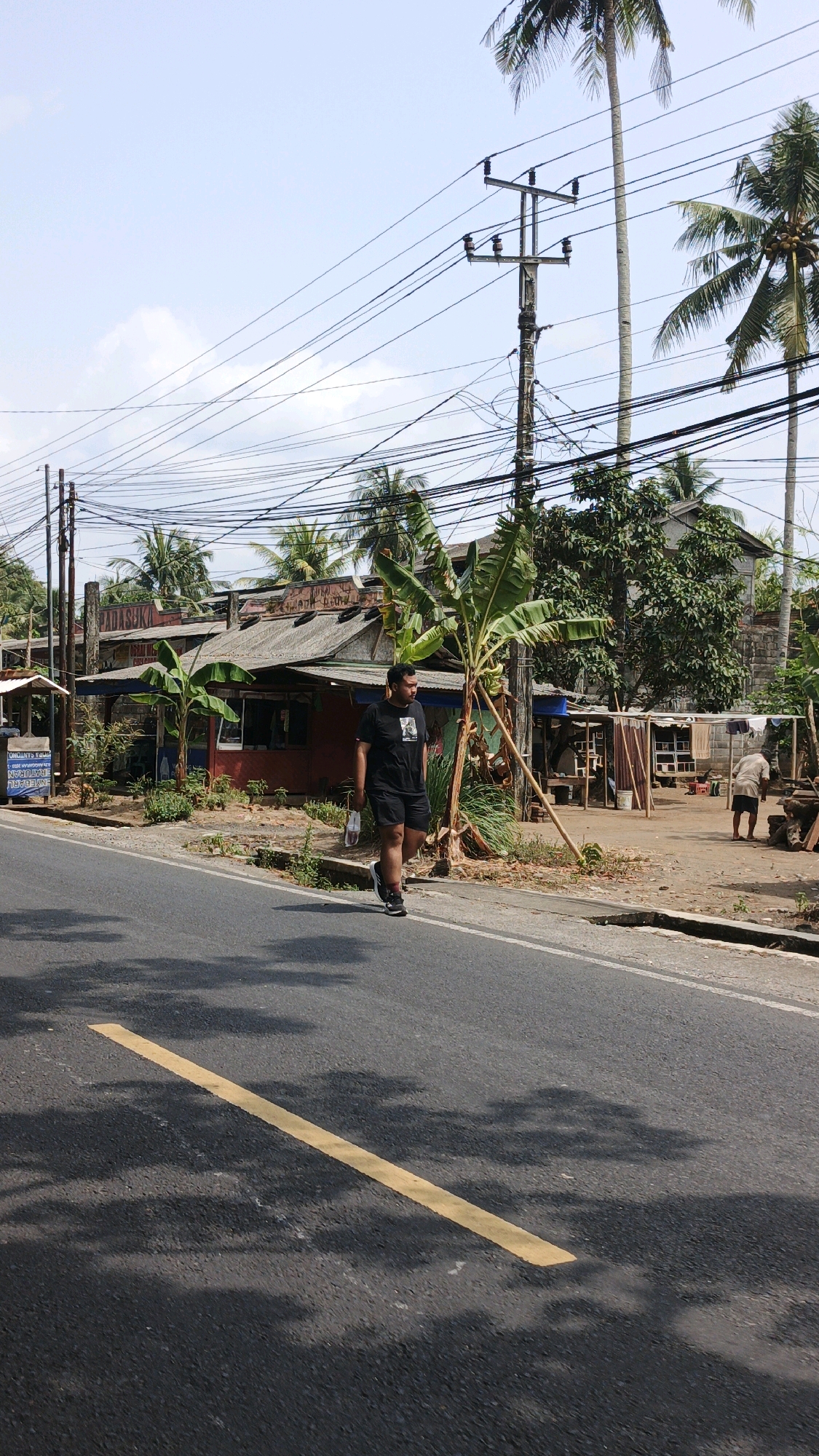 A man walks on the side of a road in front of a building with a red facade and a corrugated metal roof. The man is wearing a black t-shirt and black shorts and is carrying a white plastic bag. He is walking away from the camera towards the right side of the frame. In the background, there are utility poles with many wires, palm trees, and other vegetation. A second person, wearing light-colored clothing, is visible in the background on the right, bent over. A yellow line is painted on the road surface in the foreground. The sky is blue with scattered clouds. The scene appears to be in a semi-rural or suburban area.