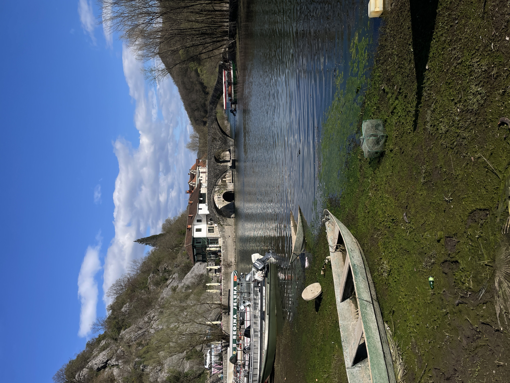 The image depicts a panoramic view of the Crnojević River and its surroundings in Rijeka Crnojevića, Montenegro, under a partially cloudy blue sky. In the immediate foreground, a muddy, moss-covered riverbank slopes gently into the water, displaying an old, light green wooden rowboat partially beached, a smaller, mostly submerged boat, a round concrete object, a green plastic fishing net, and a discarded green bottle.
The Crnojević River occupies the midground, with gentle ripples on its surface and patches of green aquatic vegetation near the banks. On the left bank, a larger, light-colored tour boat named "KRALJICA JELENA" with a green canopy and outboard motors is partially beached. Behind it, a stone embankment supports several buildings, including a prominent white structure with green window frames and a red tiled roof, appearing to be a restaurant with yellow patio umbrellas set up outside. Further downstream, the historic Old Bridge (Stari Most), a stone arch bridge, spans the river with multiple arches, connecting the two banks. Another smaller tour boat, with a red and white canopy, is docked on the right bank of the river just beyond the bridge.
The background features steep, rocky hills covered with sparse deciduous trees, many still bare, rising on both sides of the river valley. The left hillside is more rugged, while the right side presents a gentler slope covered densely with bare trees. The overall impression is that of late winter or early spring. No individuals are clearly discernible.